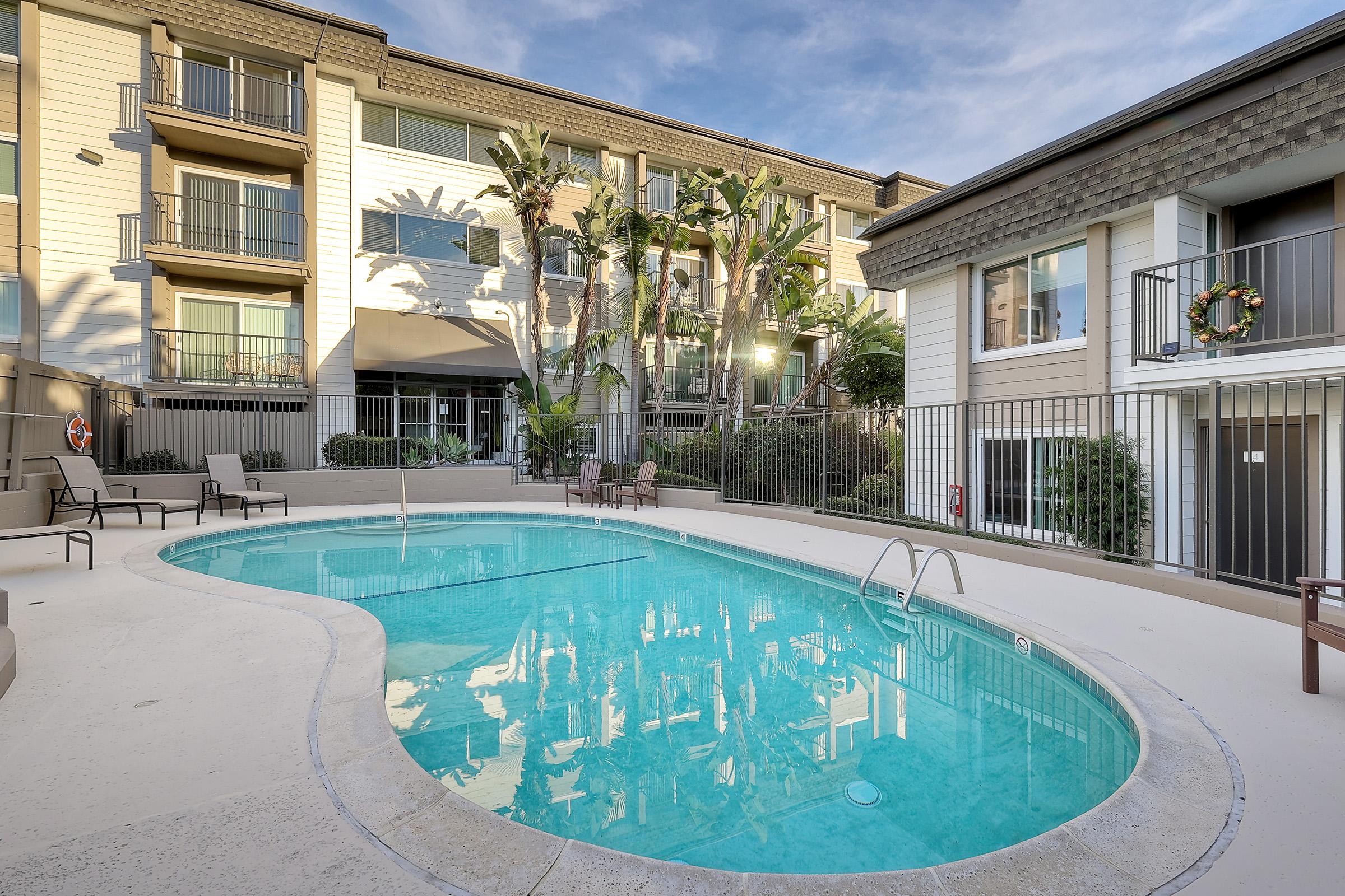 A serene outdoor swimming pool surrounded by lounge chairs, palm trees, and two multi-story building facades. The pool surface reflects the clear sky and buildings, creating a tranquil atmosphere for relaxation.