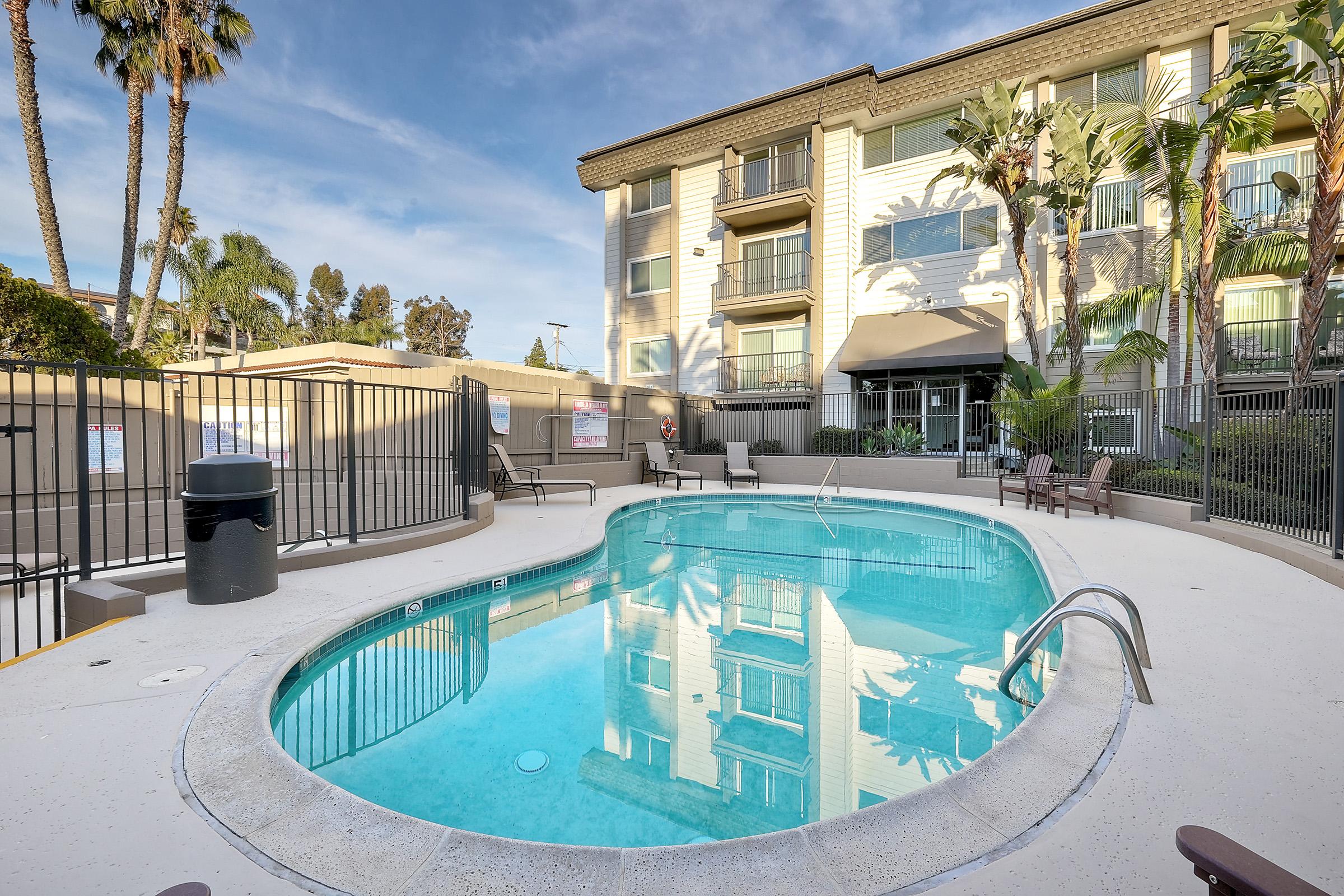 A sparkling blue swimming pool surrounded by palm trees and lounge chairs, with a modern apartment building in the background. The pool area features a gated entrance, and the overall setting is bright and inviting under a clear sky.