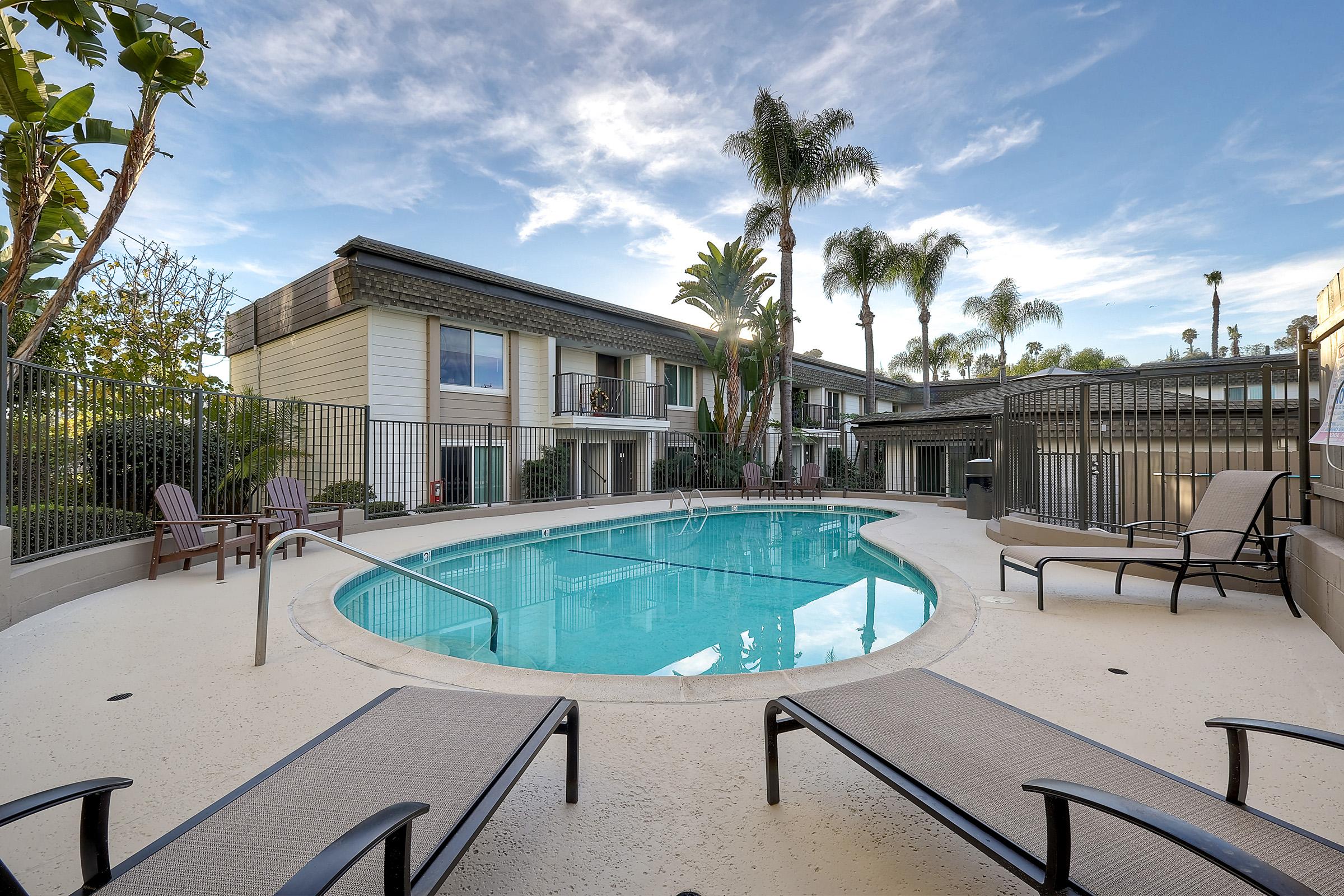 A serene pool area surrounded by palm trees and lounge chairs, with an apartment building in the background. The clear water of the pool reflects the blue sky and fluffy clouds, creating a relaxing atmosphere. Fencing encircles the pool, enhancing privacy and safety.