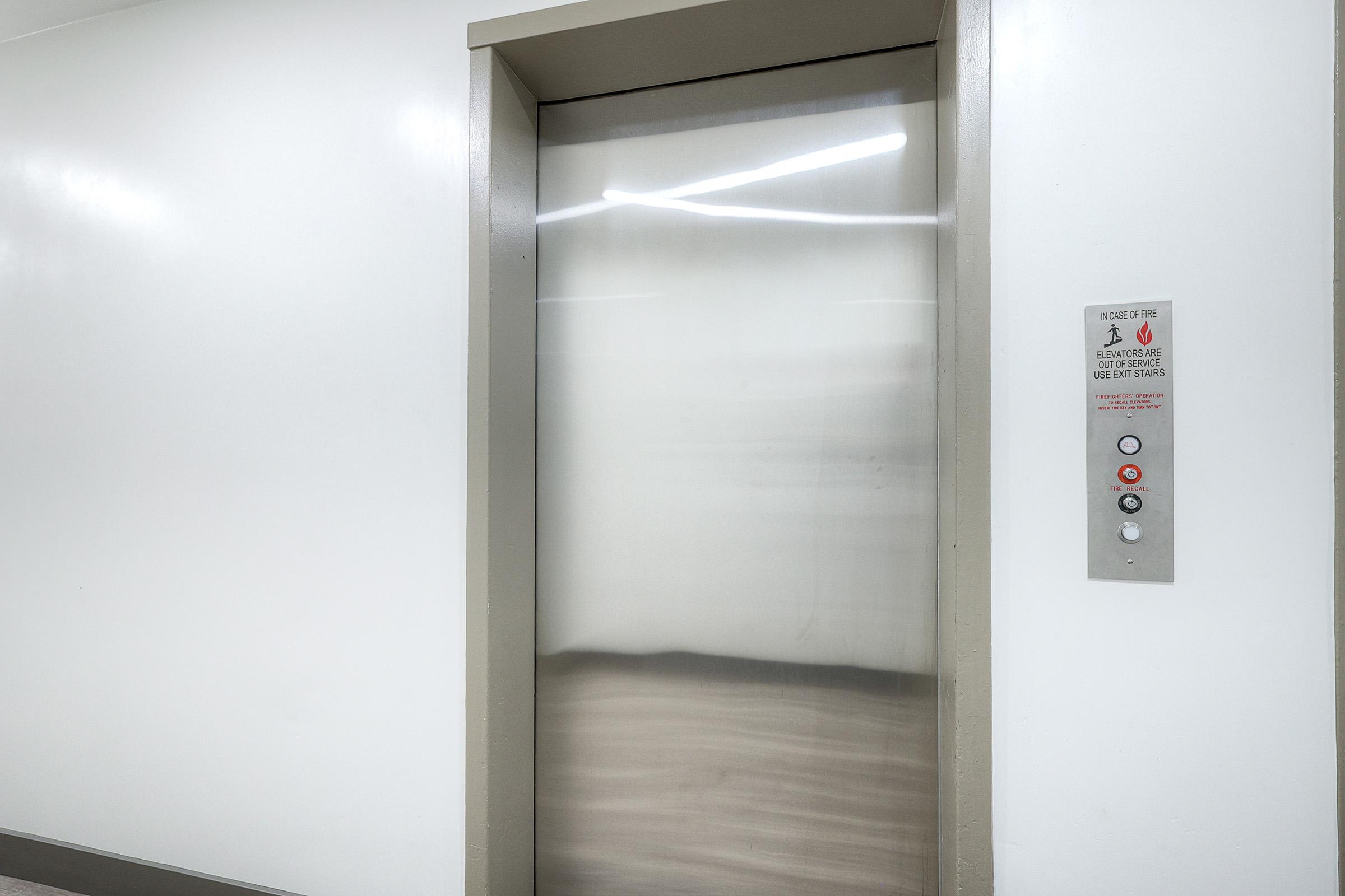 A modern elevator with a stainless steel door, located in a well-lit corridor. An elevator control panel is mounted beside the door, featuring multiple buttons for different floors. The walls are plain and white, and the floor has light wood grain texture.