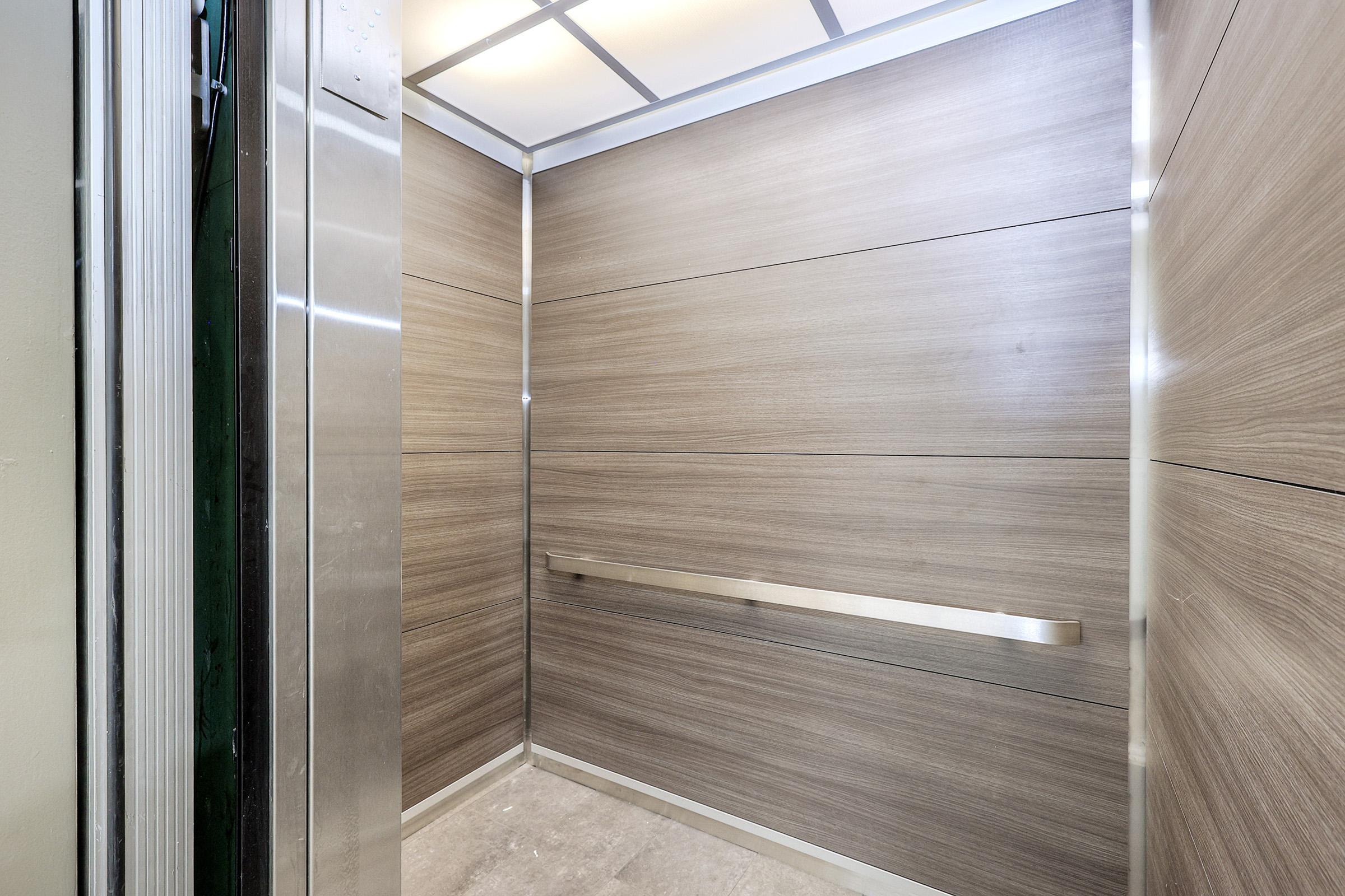 Modern elevator interior with wood paneling on the walls, smooth metallic handrail, and bright ceiling lights. The floor is made of neutral-colored tiles, creating a clean and minimalistic design.