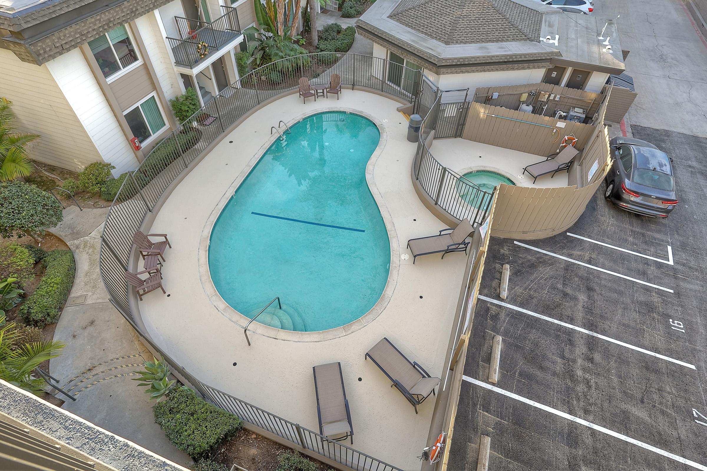 Aerial view of a swimming pool surrounded by a fence, with several lounge chairs placed around it. Nearby, there is a parking area with several spaces visible. The pool area is landscaped with greenery, while the building in the background features a light-colored exterior.