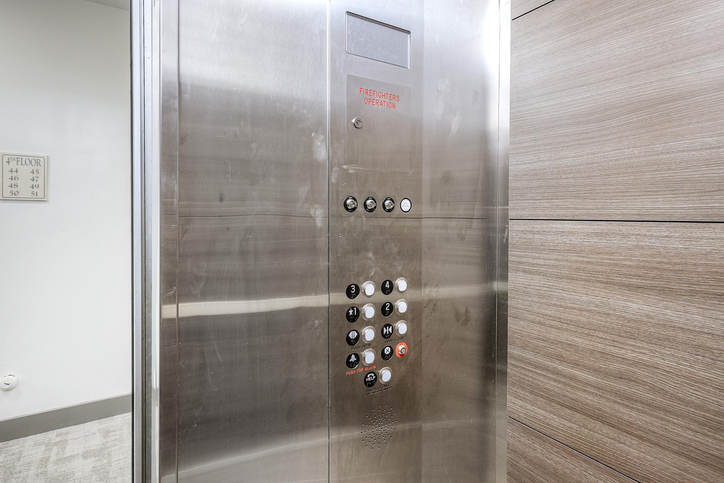 Interior of an elevator with a brushed metal panel featuring buttons for different floors. A sign above the buttons indicates "Firefighters Operate." The wall has a light wooden paneling, and there is a floor indicator showing the current floor number.