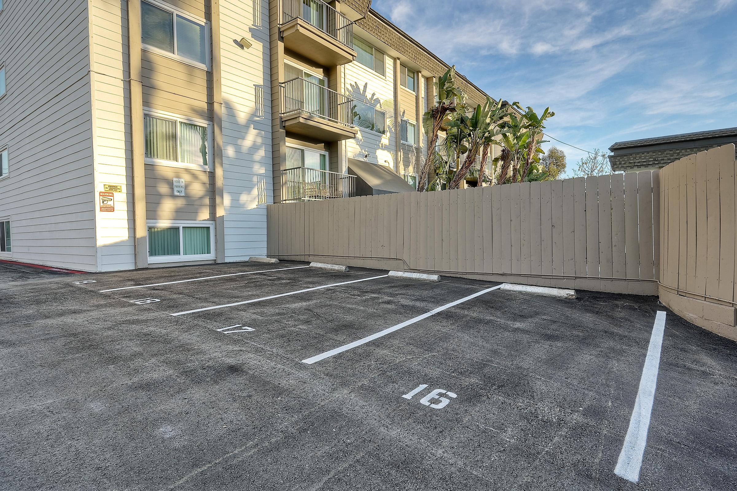 Parking lot with numbered spaces alongside a small building. The building has multiple floors and balconies, surrounded by palm trees and greenery. A wooden fence partially encloses the parking area. The sky is clear with a few clouds.