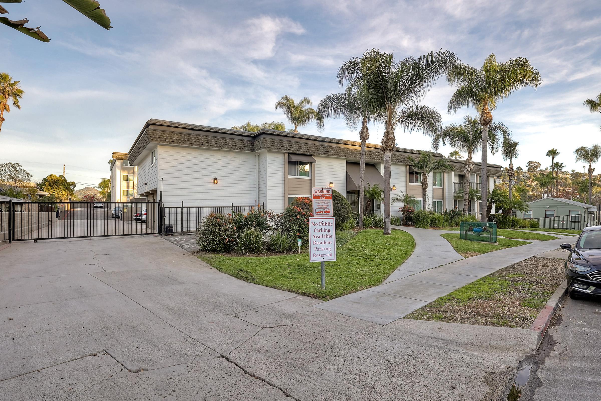 A view of a two-story building surrounded by palm trees and landscaping. There is a gated entrance with a sign indicating no public access. A curving pathway leads to the entrance, and parking spaces are visible. The scene is set in a sunny environment with clear skies.