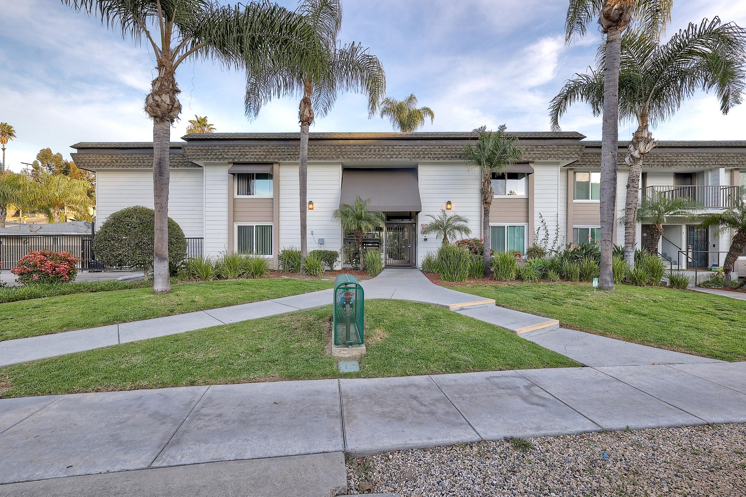 A landscaped walkway leads to the entrance of a modern apartment building with a flat roof. Palm trees and shrubs frame the entrance, which features large glass doors and a welcoming atmosphere. Paths of concrete curve through green grass, providing access to the front door and surrounding gardens.