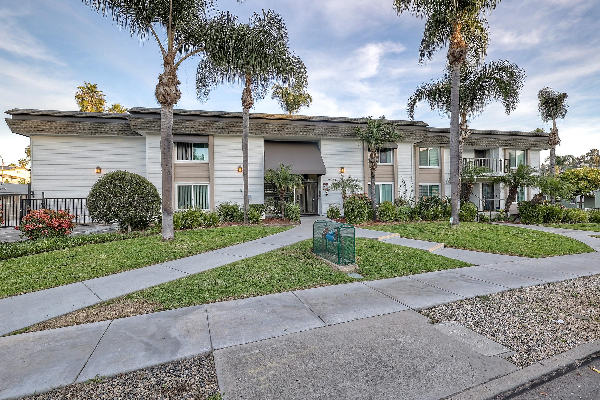 A modern apartment complex surrounded by lush greenery and palm trees. The building features a central entrance with large windows and landscaped grounds with manicured lawns and flower beds. A pathway leads from the sidewalk to the entrance, creating an inviting atmosphere.