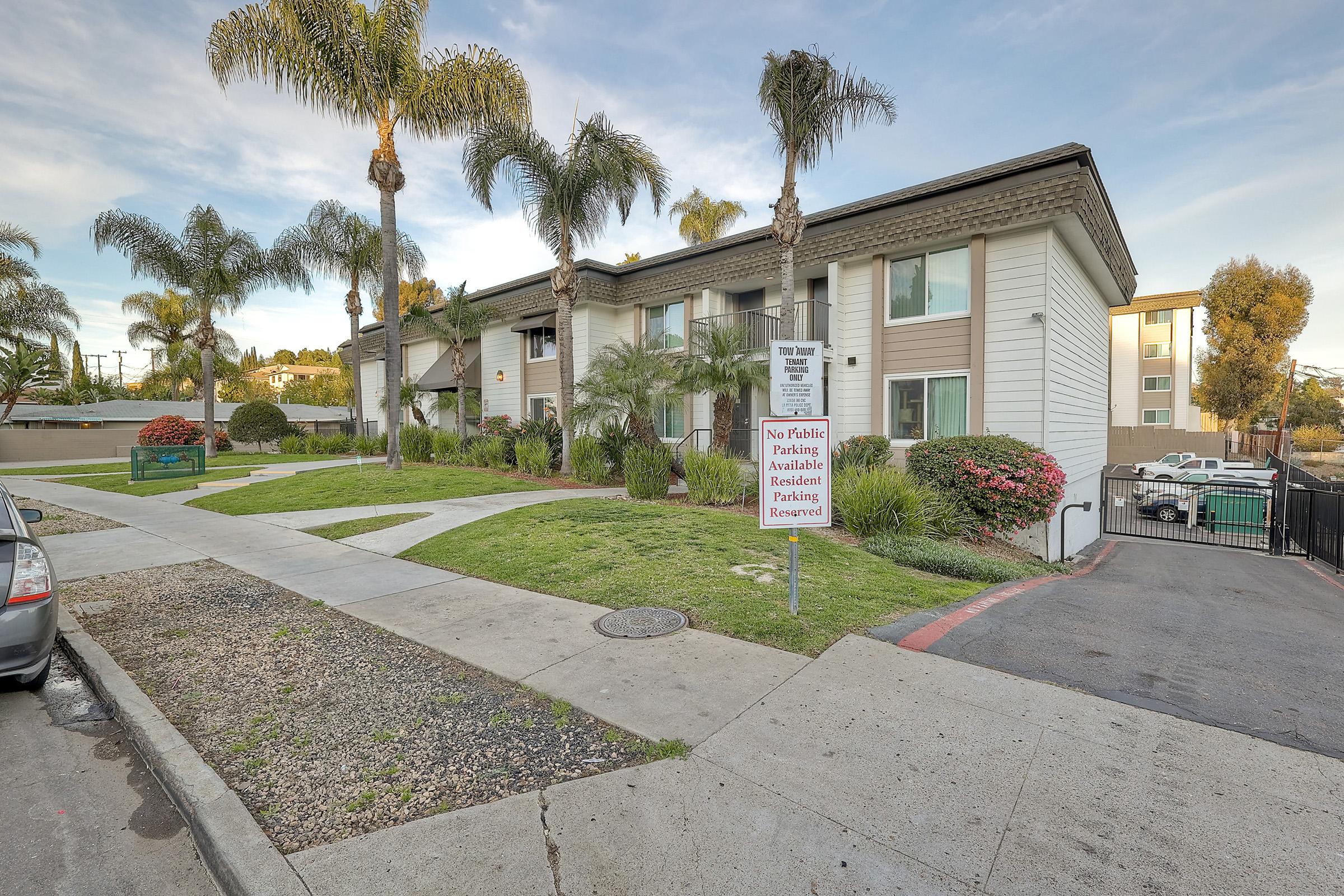 A small apartment building with a white exterior and brown accents, surrounded by green lawns and palm trees. A sign indicates no public parking is available. Flowering bushes add color to the landscape. A parking area is visible in the foreground, along with a vehicle parked near the curb.