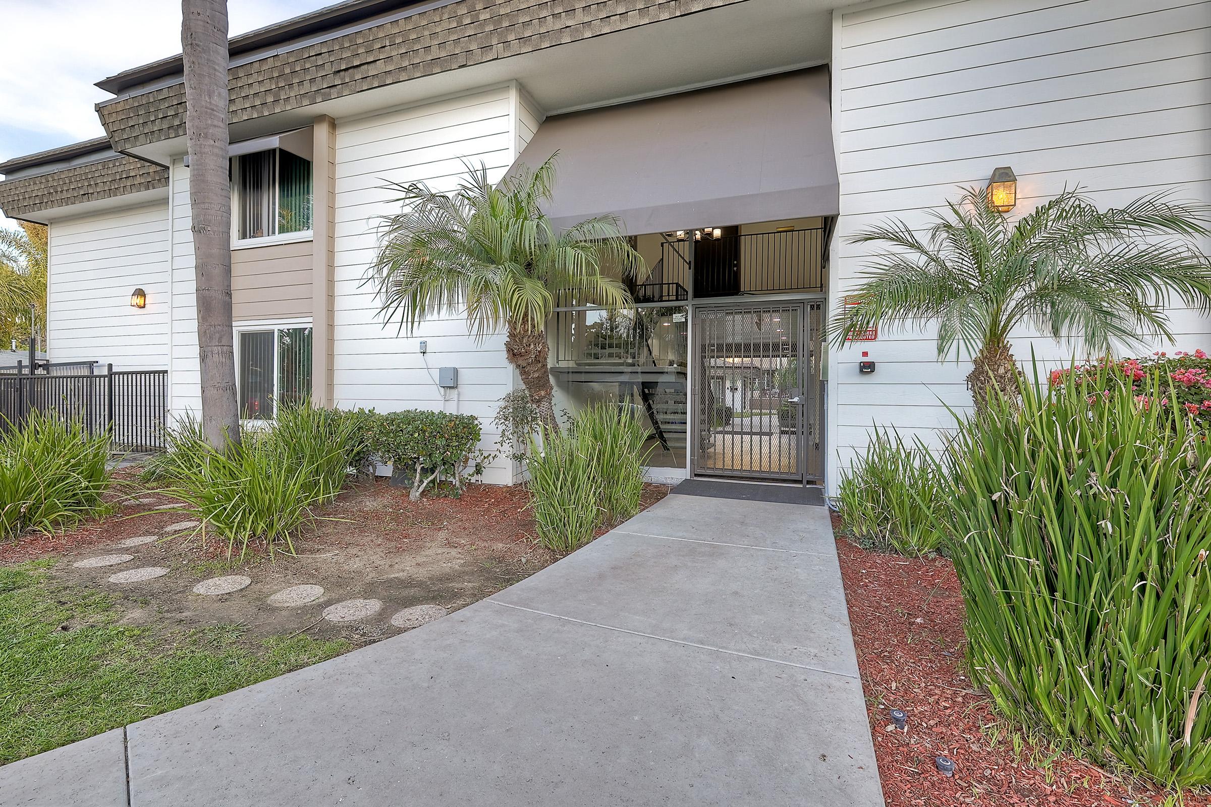 Entrance of a modern multi-story building with a palm tree and bushes in the foreground. A concrete pathway leads to the entrance, which features large glass doors and a stairway visible inside. The surroundings are landscaped with red mulch and green plants.