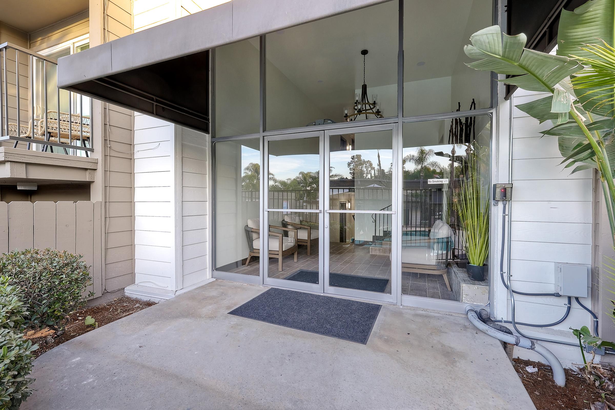 Entrance to a modern building featuring large glass doors, a basic mat at the threshold, and landscaped greenery on either side. Inside, visible furniture and decor hint at a welcoming lobby area, with a chandelier hanging from a high ceiling. The setting includes a patio or garden view beyond the doors.
