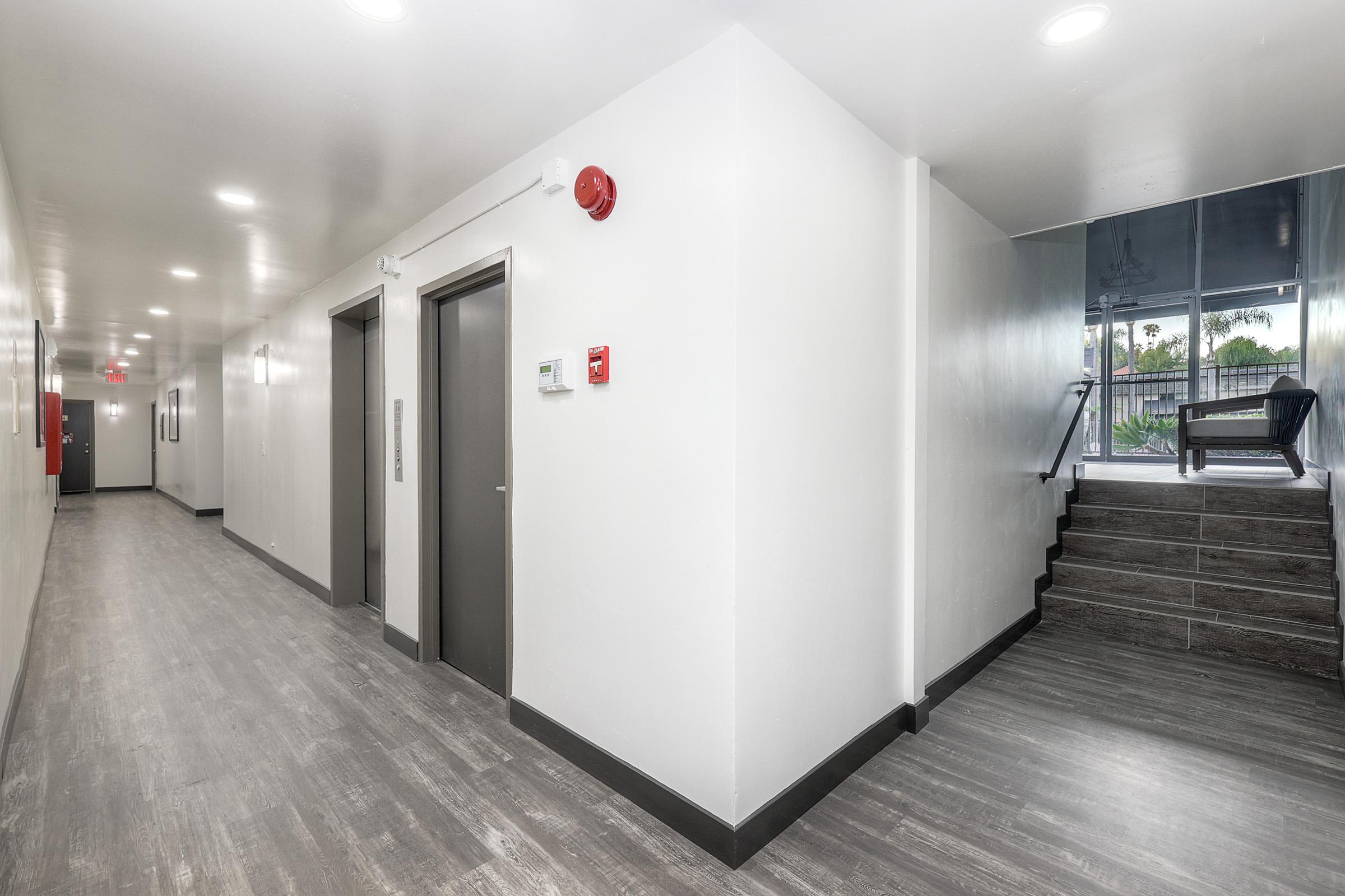 A well-lit, modern indoor hallway featuring smooth gray flooring, elevators on the left, fire alarm and emergency exit signage, and a staircase leading to a glass door at the end, which opens to an outdoor area with seating.