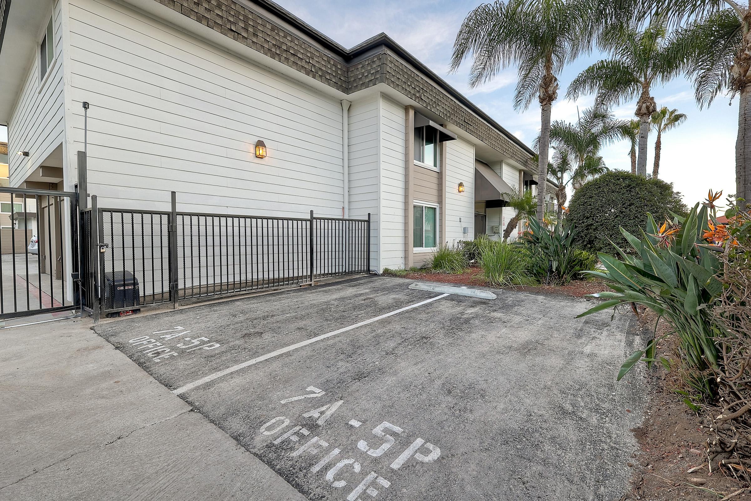 A view of a small parking area in front of a modern white building with palm trees and shrubs. A fence surrounds the parking spaces, which are marked "7A-5P OFFICE." The setting is well-maintained and features Mediterranean-style landscaping.