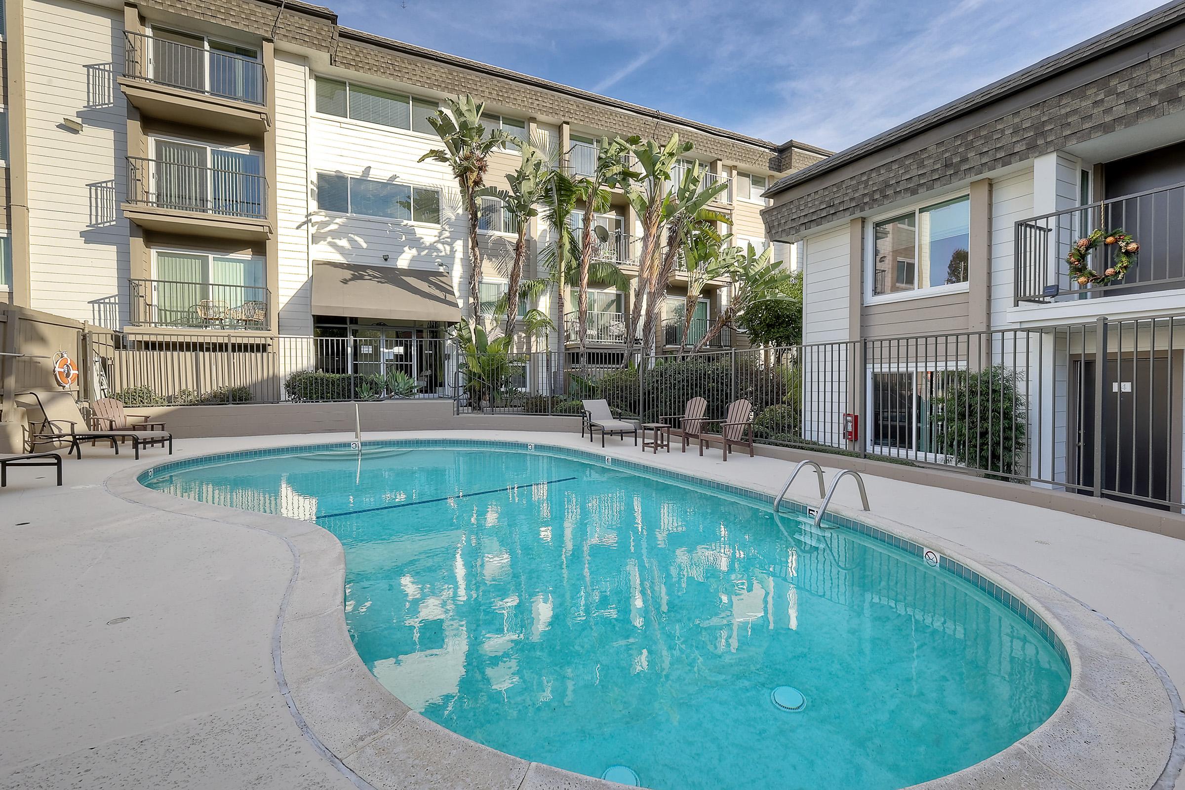A serene swimming pool surrounded by palm trees and lounge chairs, located in a courtyard area of an apartment complex. The multi-story building features balconies and large windows, with a bright blue sky in the background.