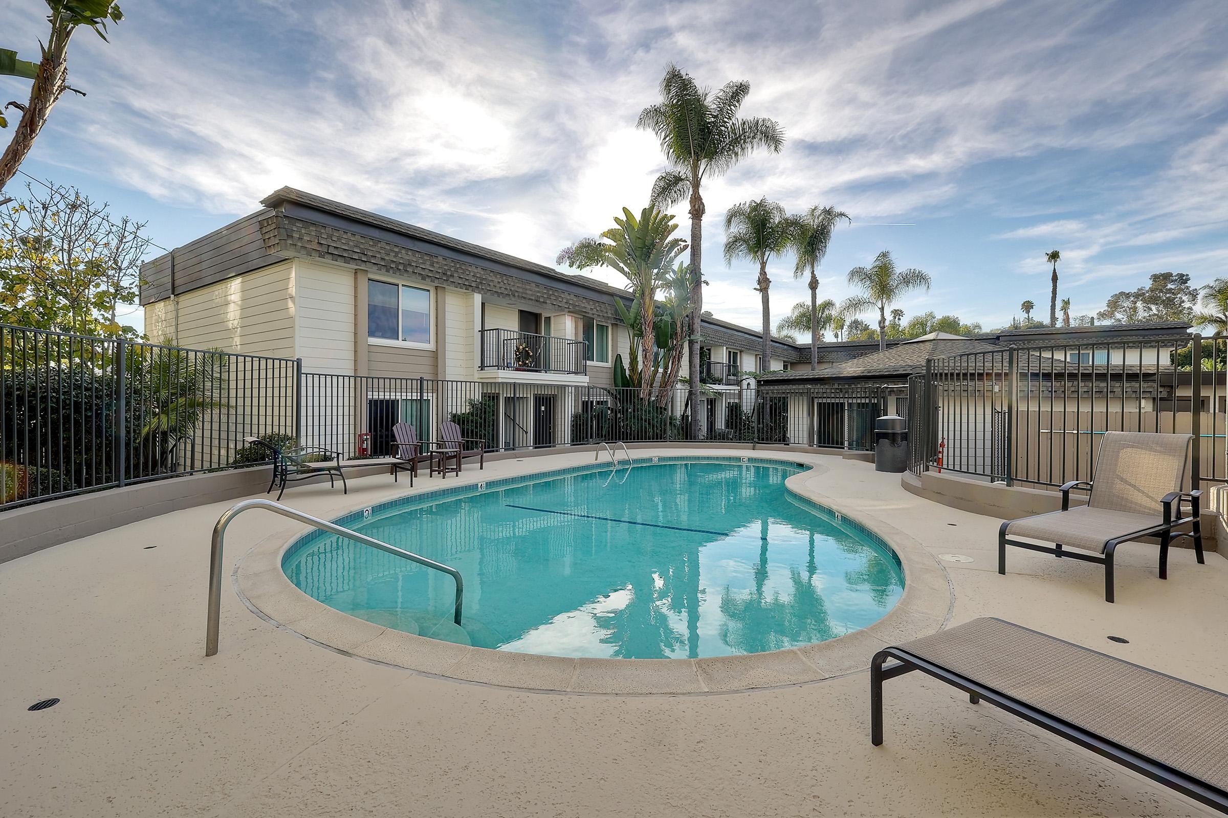 A serene pool area surrounded by palm trees with sunlit apartments in the background. The pool is circular with a textured deck and lounge chairs nearby, set against a clear blue sky. The atmosphere is inviting and tranquil, perfect for relaxation.