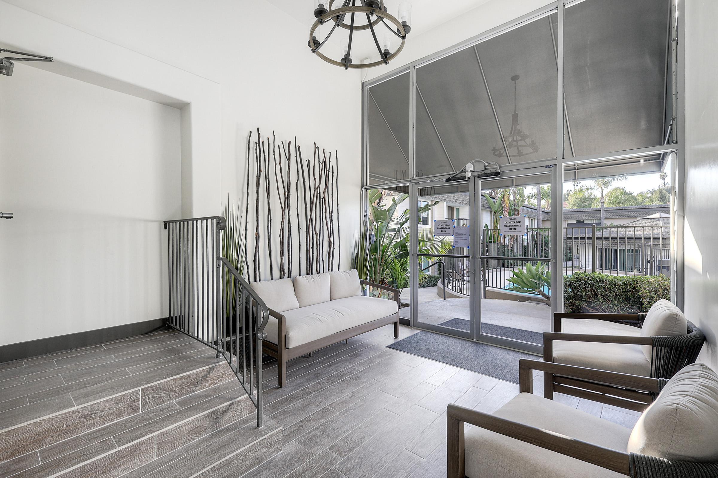 A modern entryway featuring a minimalist design with a chandelier, two wooden-framed chairs, and a sofa. The space is enhanced by tall decorative branches on the wall and large windows that open to a patio area surrounded by greenery.