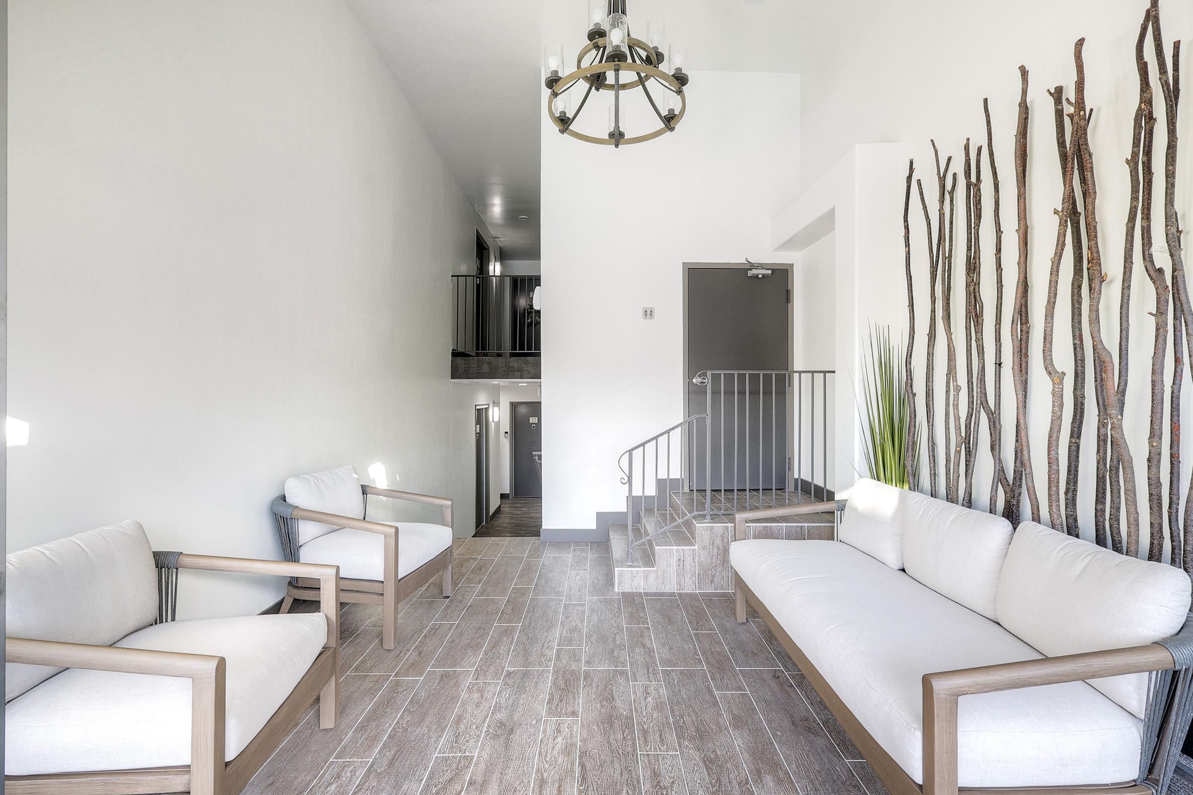 Bright and modern interior of a lobby or waiting area featuring two light-colored sofas with wooden frames. A staircase leads to an upper level in the background, while a decorative arrangement of tall, slender branches is placed against the wall. A chandelier hangs from the ceiling, adding a touch of elegance.