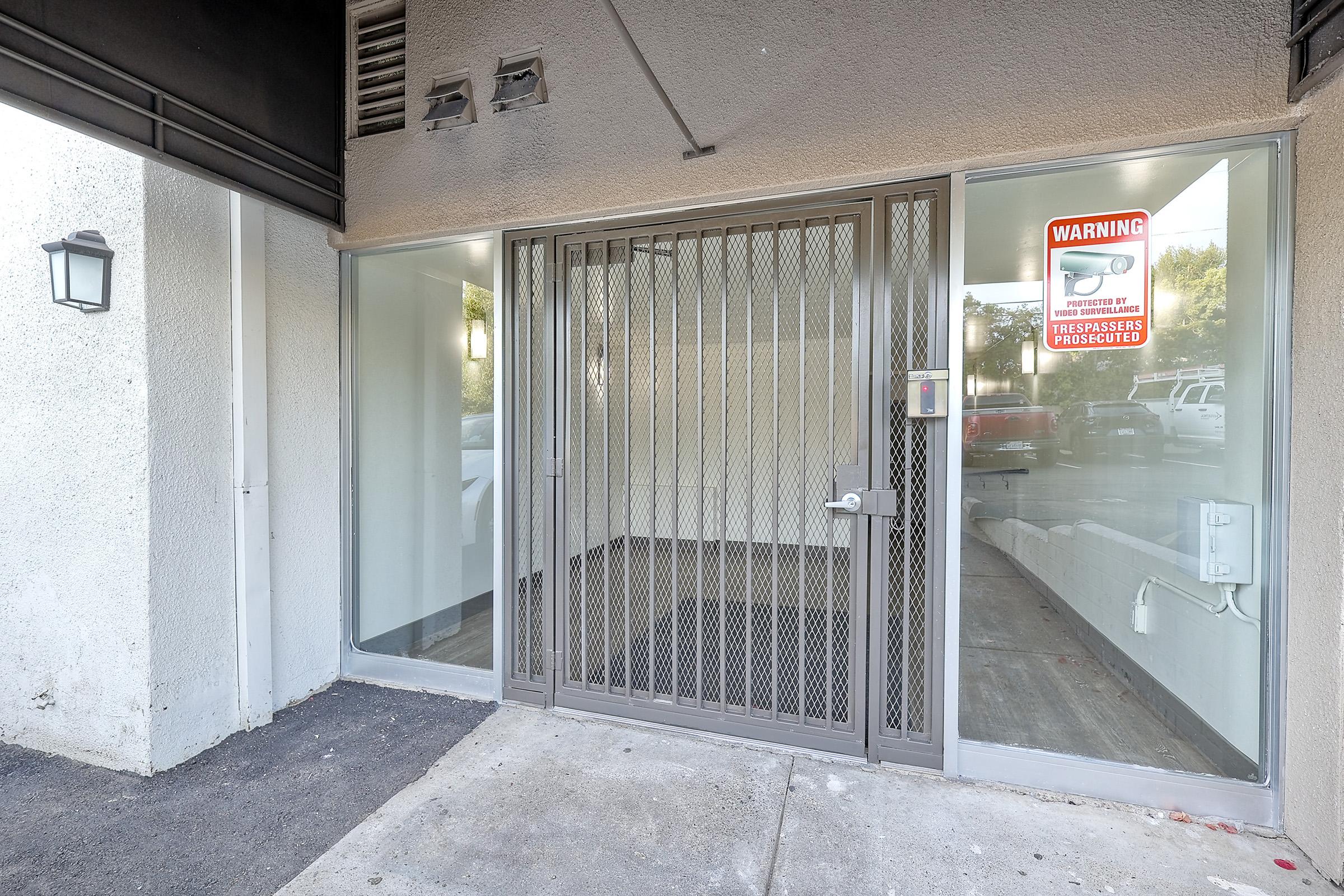 A closed entrance with a metal security gate and glass windows on either side. A sign on one window displays a warning, and the door is locked. The area has a concrete surface and is well-lit, with a light fixture mounted on the wall.