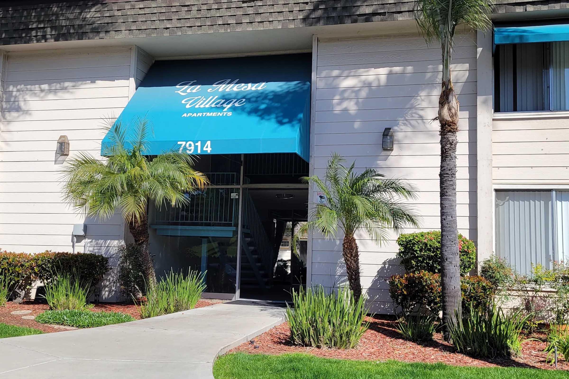 Entrance to La Mesa Village Apartments, featuring a blue awning with white text. The pathway leads to the doorway, flanked by palm trees and shrubs. Bright daylight illuminates the scene, highlighting the building's exterior and landscaping.