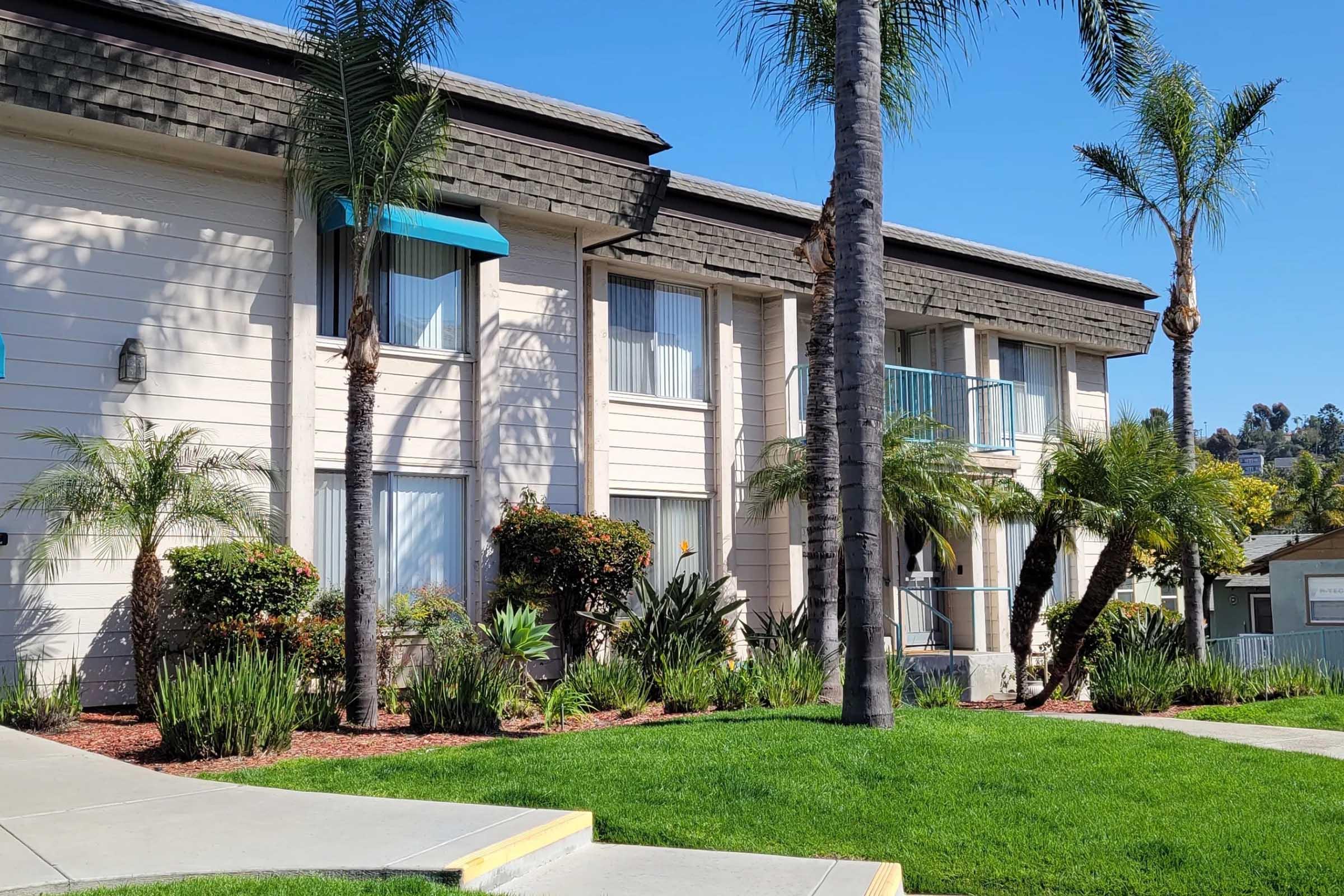 A light-colored apartment building featuring three stories, palm trees, and well-maintained landscaping with shrubs and grass. The building has balconies, large windows, and a clear blue sky in the background.