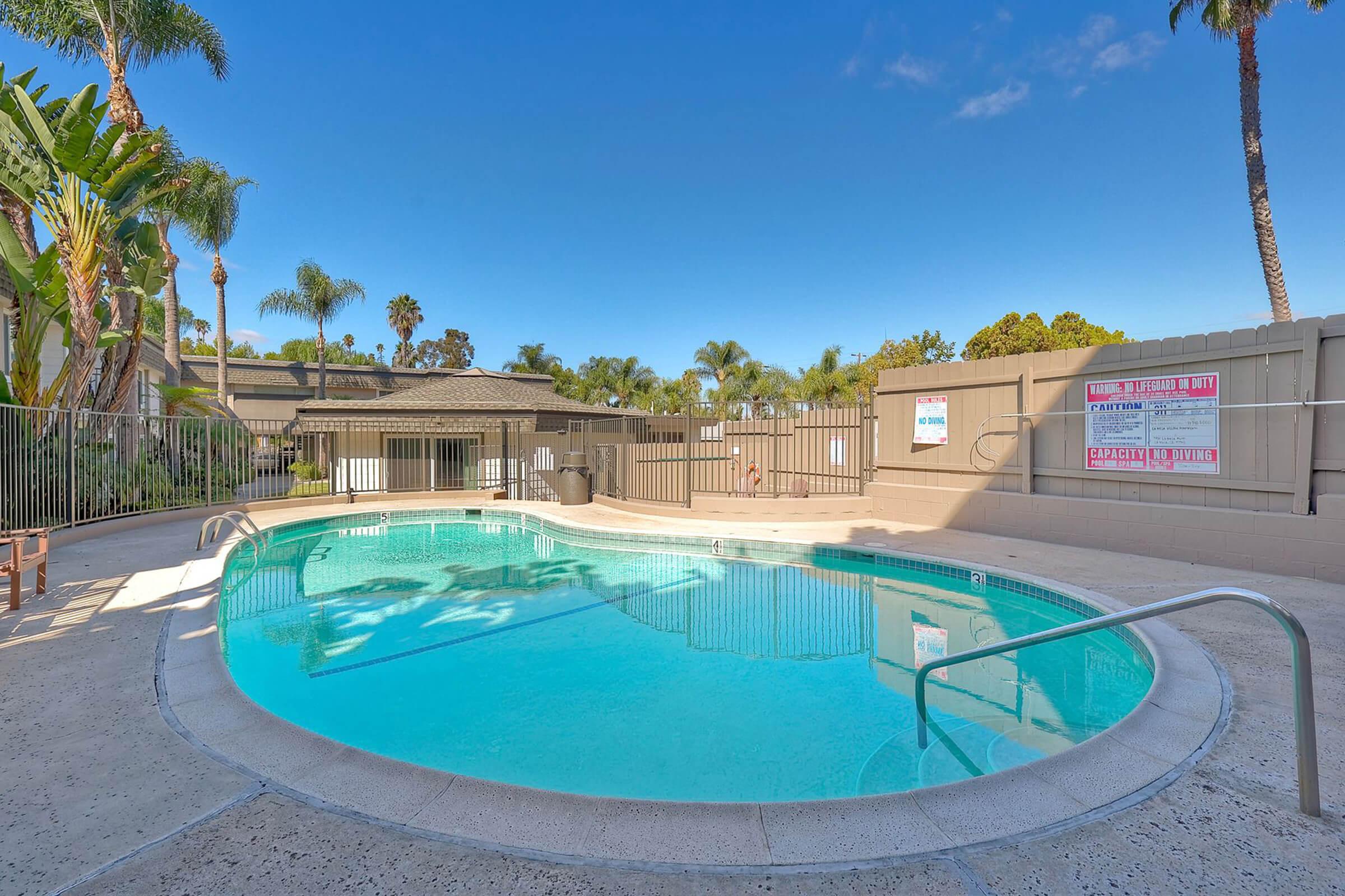 A clear swimming pool surrounded by a tiled deck and green palm trees, under a blue sky. A fence encloses the area, and there are signs on the walls nearby. The pool features a shallow entry and is free of people, creating a peaceful, inviting atmosphere.