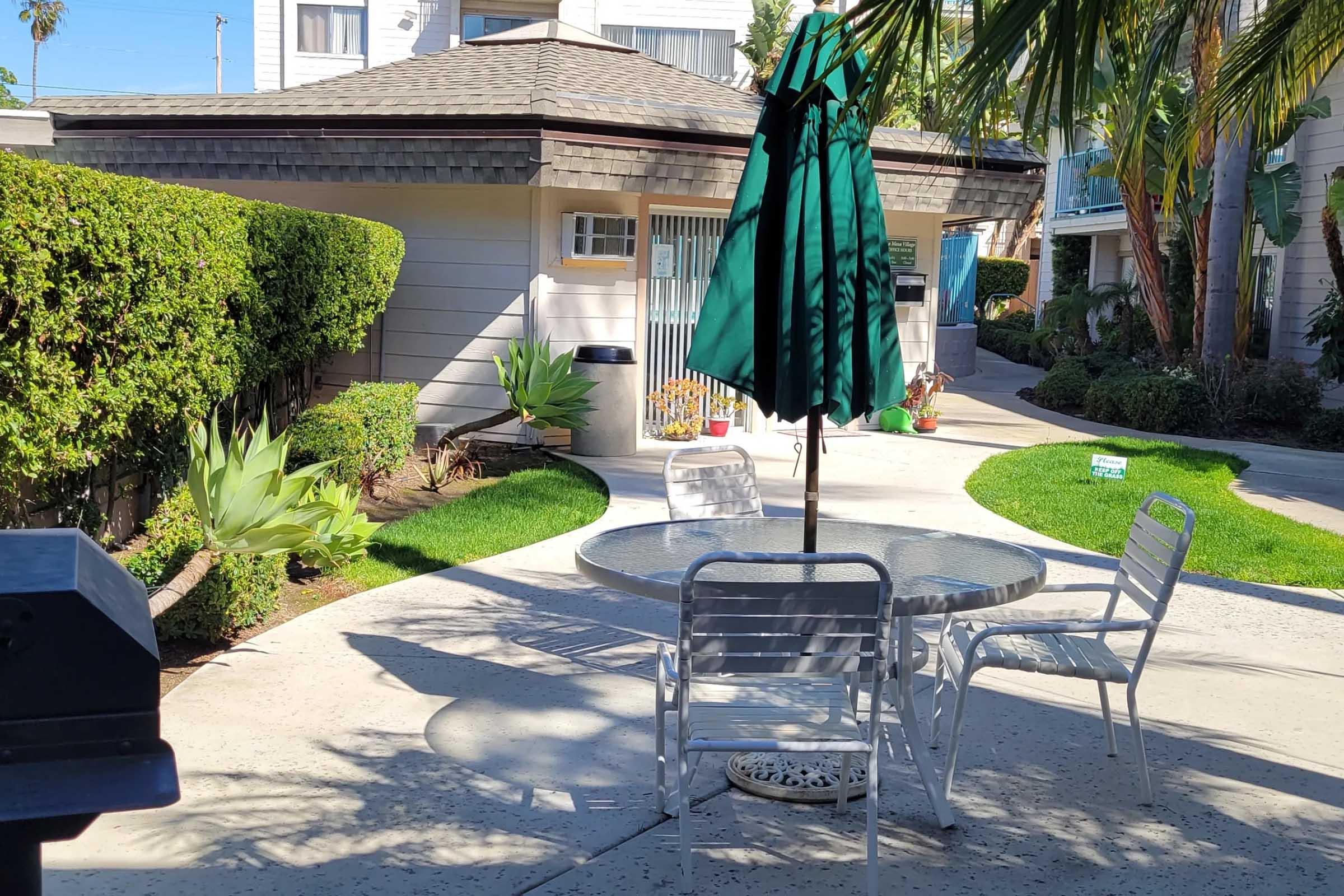 A patio area with a round table and four metal chairs under a green umbrella. Lush greenery and shrubs surround the space, and a concrete path leads to a building in the background. Bright sunlight casts shadows on the ground, creating a relaxed outdoor atmosphere.