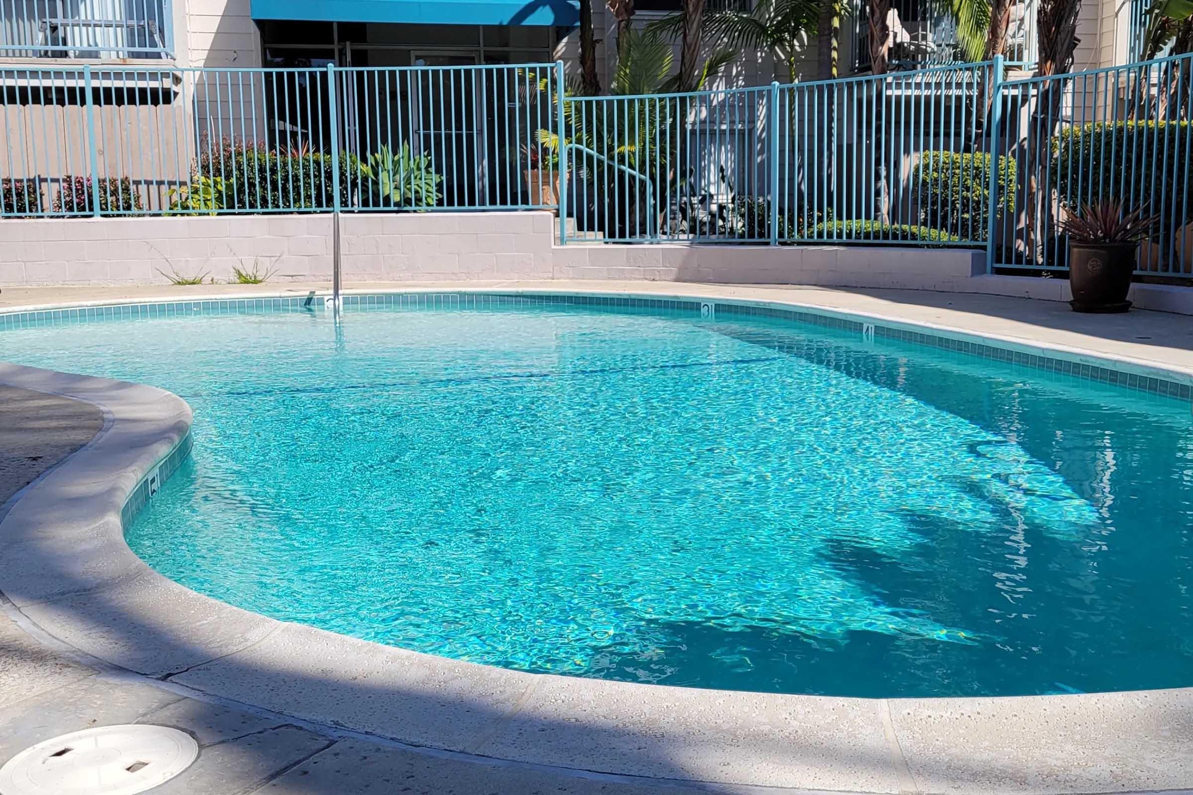 A clear swimming pool with sparkling turquoise water, surrounded by a fence and lush greenery. Sunlight reflects on the surface of the water, creating a serene and inviting atmosphere. The area is well-maintained, featuring a paved border around the pool.