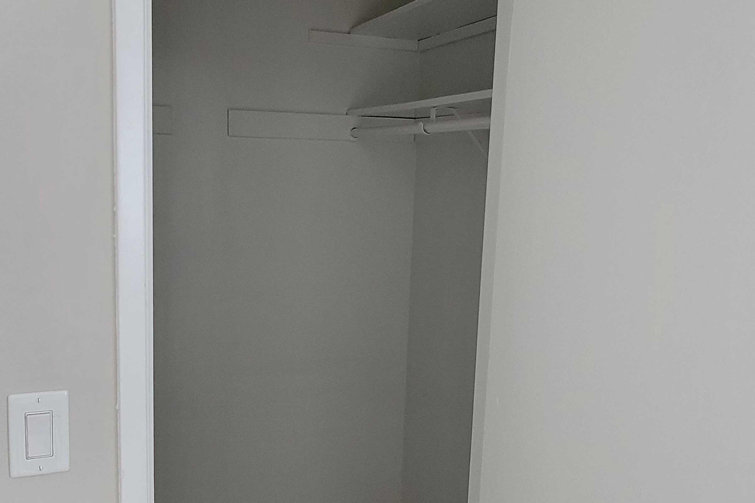 A minimalist empty closet with white walls and a shelf on top. There are two horizontal hanging rods visible, and the floor is bare. The closet is well-lit, creating a clean and spacious appearance.