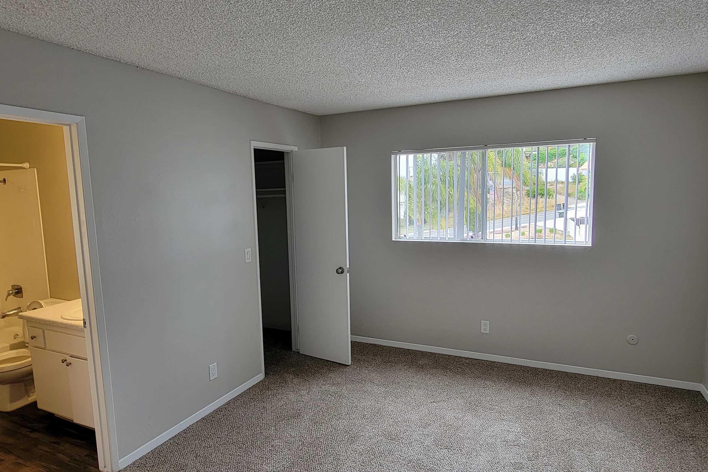 A bright, empty room with light gray walls and carpeted flooring. There is a window with blinds letting in natural light. A door leads to a bathroom, and another door is slightly ajar, revealing a closet. The space appears freshly painted and ready for furnishings.