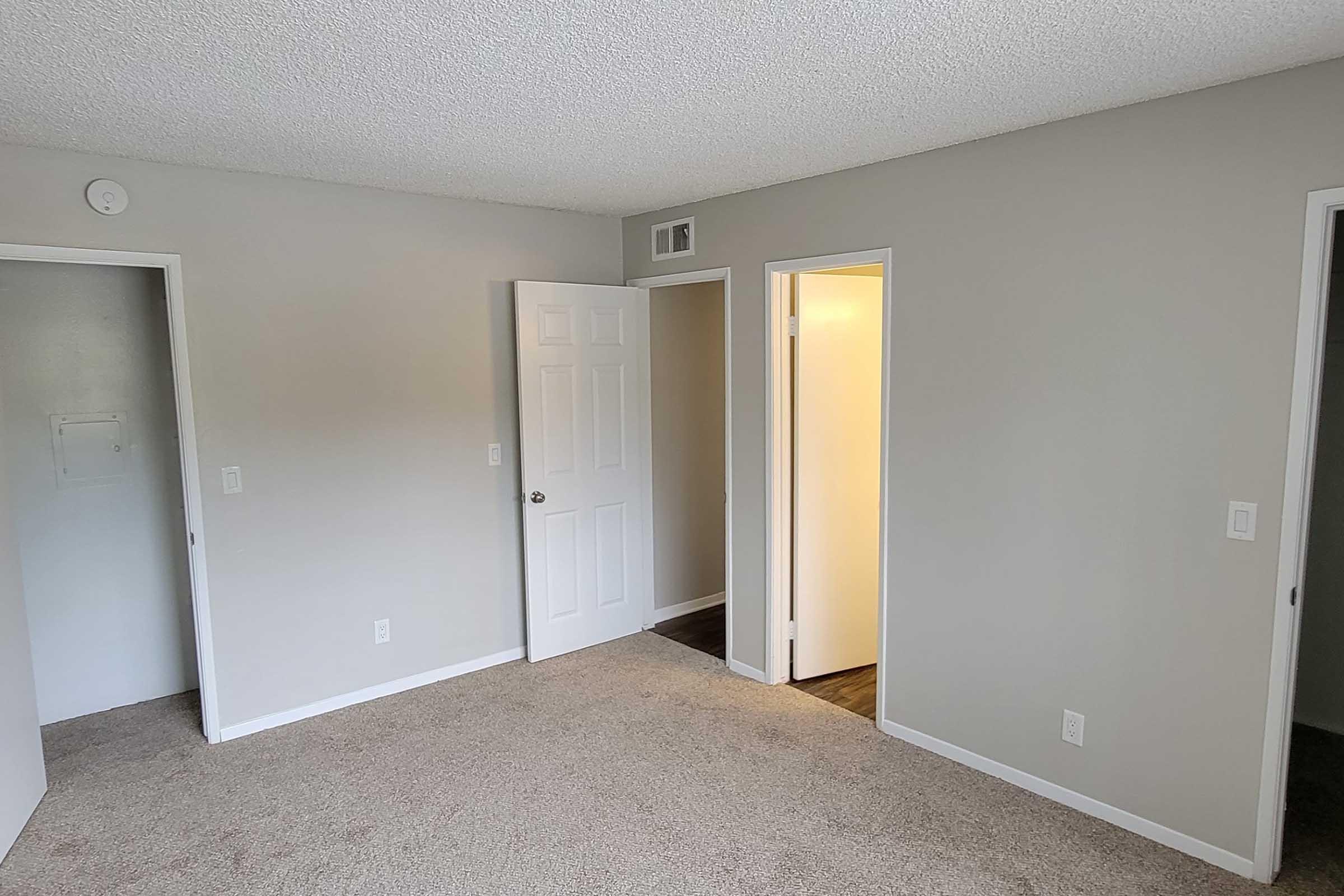 A neutral-colored room with textured walls and carpeted flooring. There are two doorways, one leading to a closet and the other to a bathroom or hallway. Natural light enters from an open door, creating a bright, inviting atmosphere.