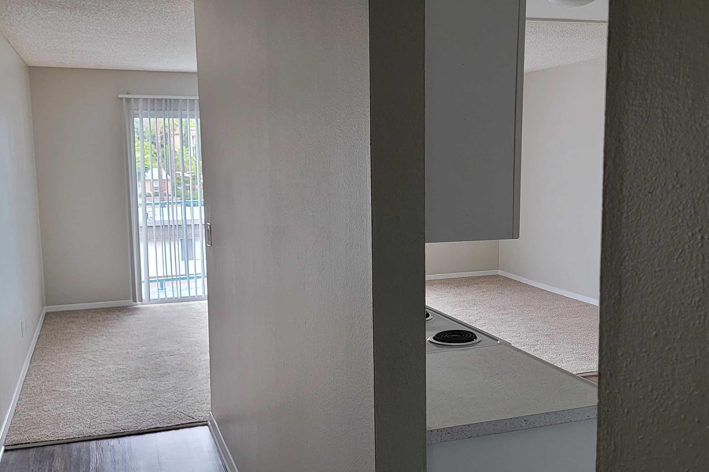 View of a small apartment interior showcasing a carpeted living area and a kitchen space. There is a window with blinds allowing natural light, and the walls are painted in neutral tones. The kitchen features a countertop with a stovetop, while the living area is empty, suggesting a clean and simple layout.