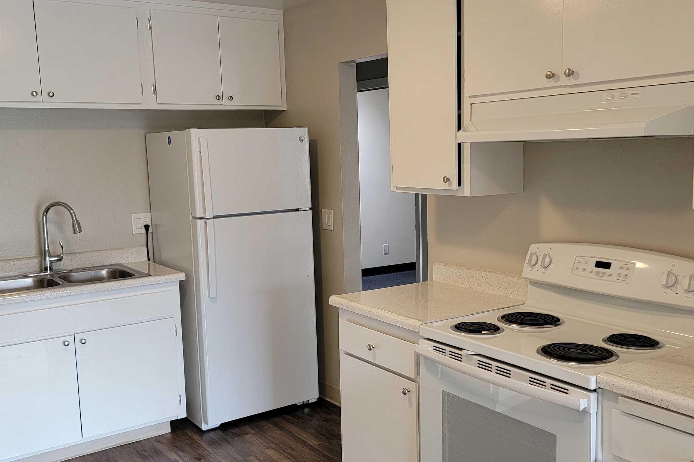 A tidy kitchen featuring white cabinets, a refrigerator, a double sink, and a stove with an oven. The countertops are light-colored, and the floor is dark wood. There is an open doorway leading to another room, indicating a spacious and functional layout.