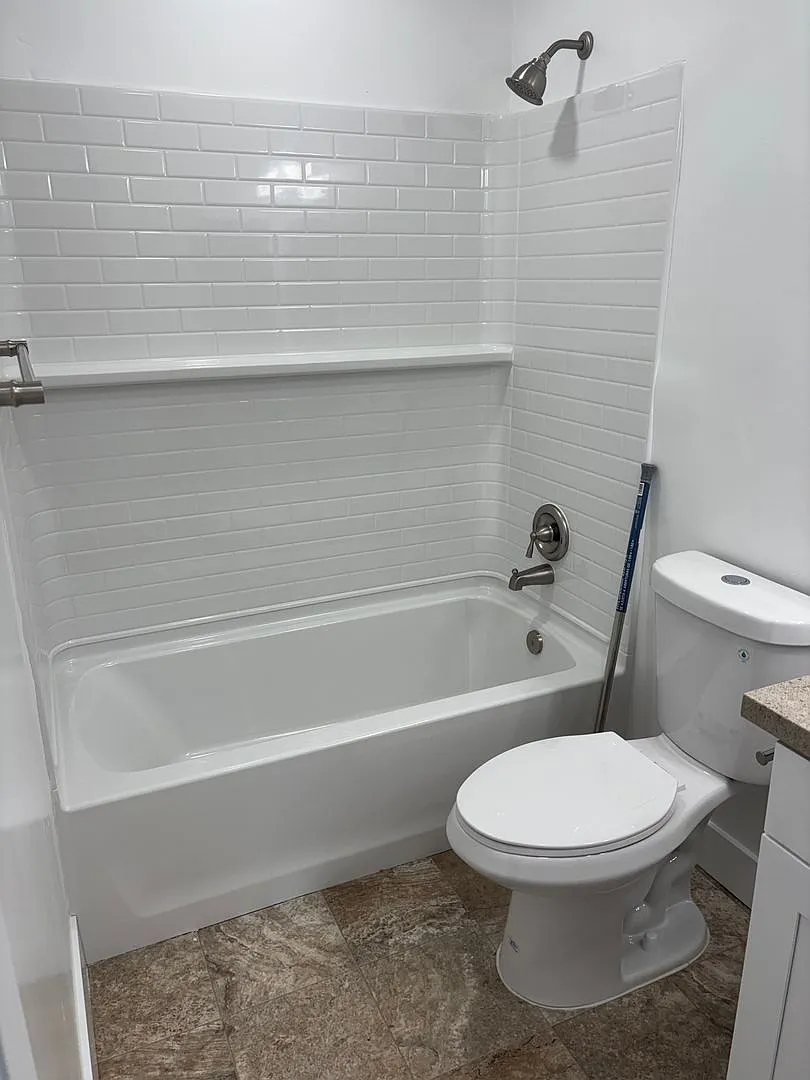 A clean, modern bathroom featuring a white bathtub with a showerhead, white subway tile on the walls, a toilet with a flush handle, and a small countertop beside it. The floor has a light-colored stone finish, creating a bright and fresh atmosphere.