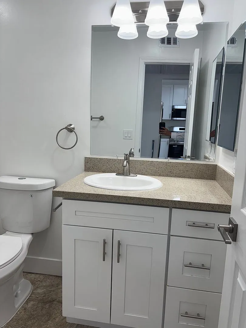 A modern bathroom featuring a white vanity with a sink, a large mirror above, a simple light fixture with three pendant lights, a toilet, and a neutral tiled floor. The space is well-lit and organized, with cabinets and drawers for storage.