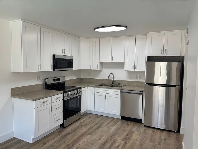 Modern kitchen featuring white cabinetry, stainless steel appliances including a microwave, oven, and refrigerator, and a sleek sink. The countertop is light-colored with a natural finish, and the floor is made of light wood laminate. A circular ceiling light provides bright illumination.
