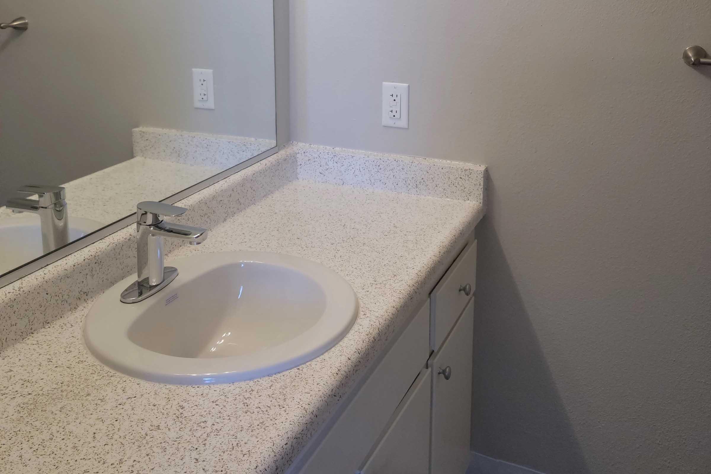 A clean bathroom countertop with a small white sink and modern faucet. The surface is light-colored with a speckled design. A large mirror hangs above the sink, and light switches are visible on the wall. The walls are painted a neutral color, and there is a towel bar on the side.
