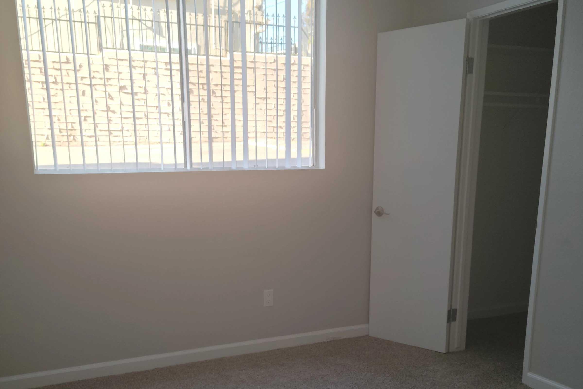 A corner of a room featuring a beige carpet and light-colored walls. There is an open white door leading to a closet, and a large window with vertical blinds allowing natural light to enter. Outside the window, a stone wall is visible. The overall atmosphere is calm and minimalistic.