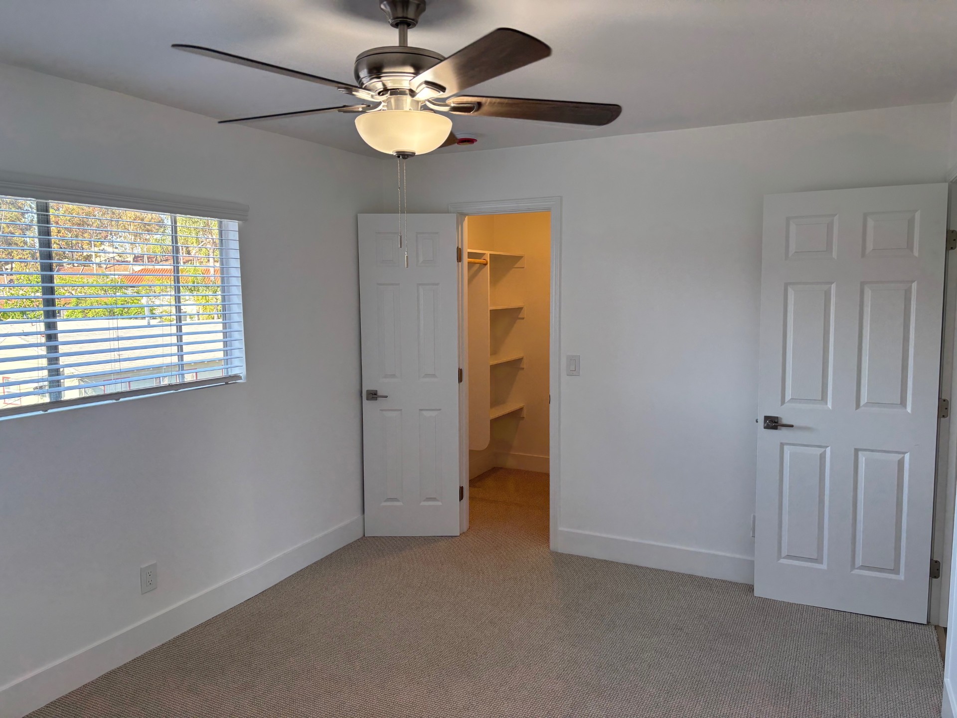 A bright, empty room featuring a ceiling fan, light fixture, and two doors—one leading to a closet—against a backdrop of white walls and carpeted floor. A window with blinds allows natural light to enter, illuminating the space.