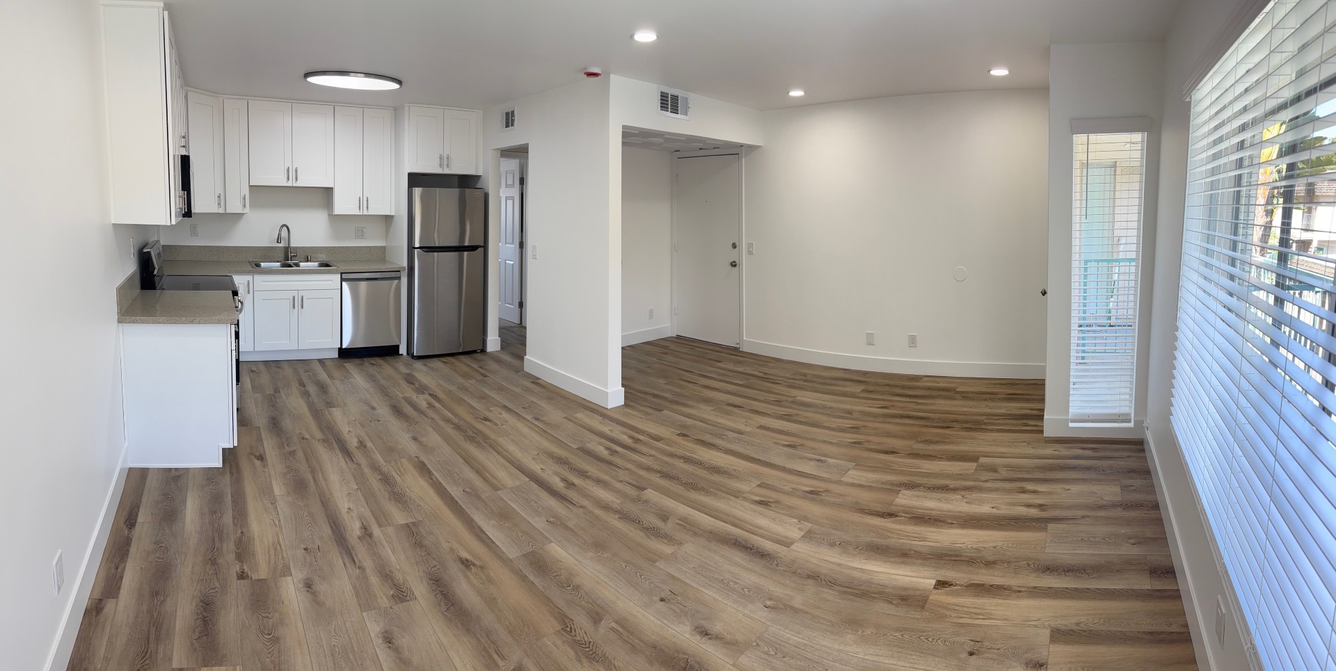 Bright and modern apartment interior featuring a spacious open layout. The kitchen, equipped with stainless steel appliances and white cabinetry, is adjacent to the living area, which has large windows letting in natural light. The flooring is light wood, enhancing the airy feel of the space.