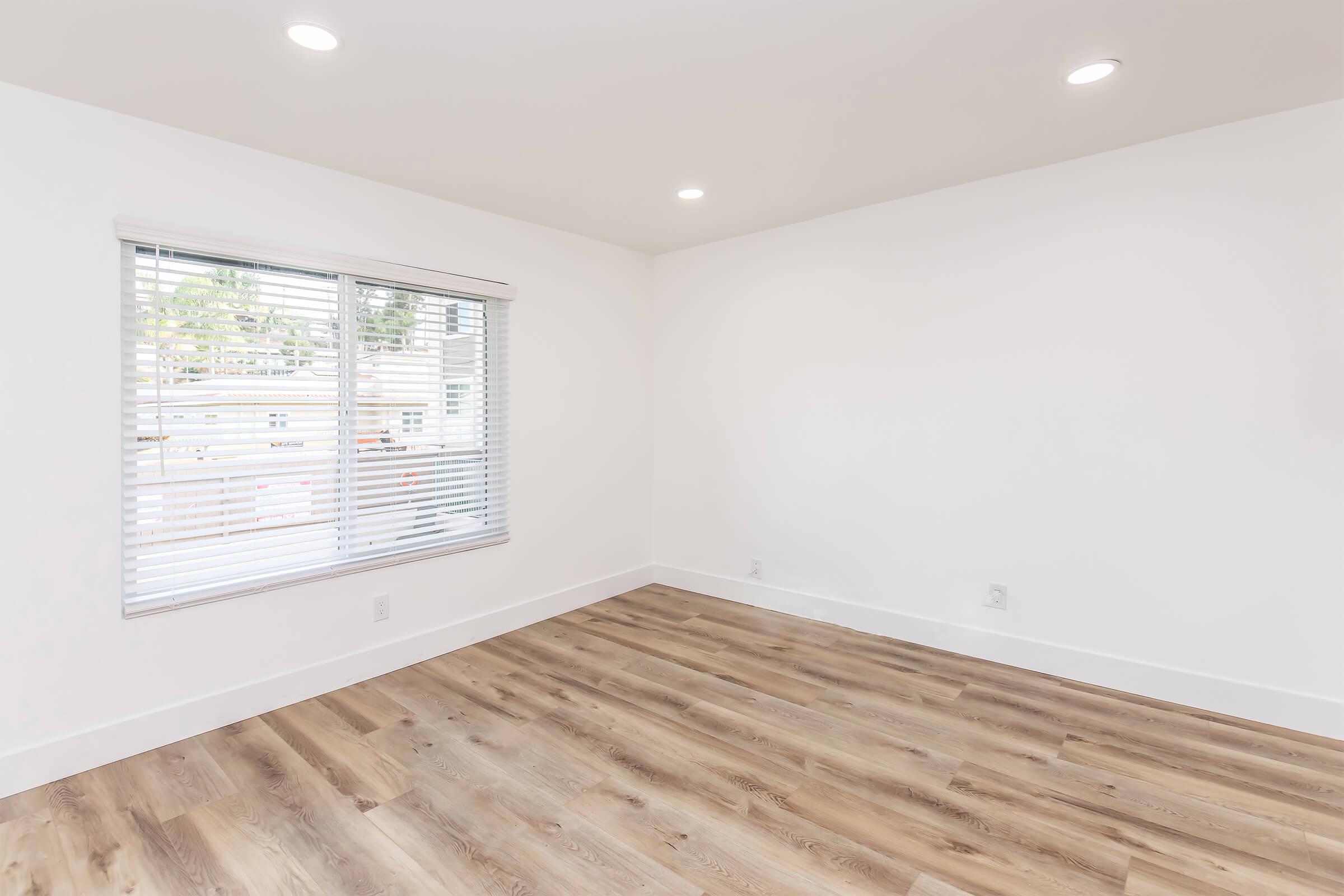 A bright, empty room featuring light-colored walls and wooden flooring. There is a large window with blinds allowing natural light to enter, creating an open and airy atmosphere. The space is minimalist, with no furniture or decorations.