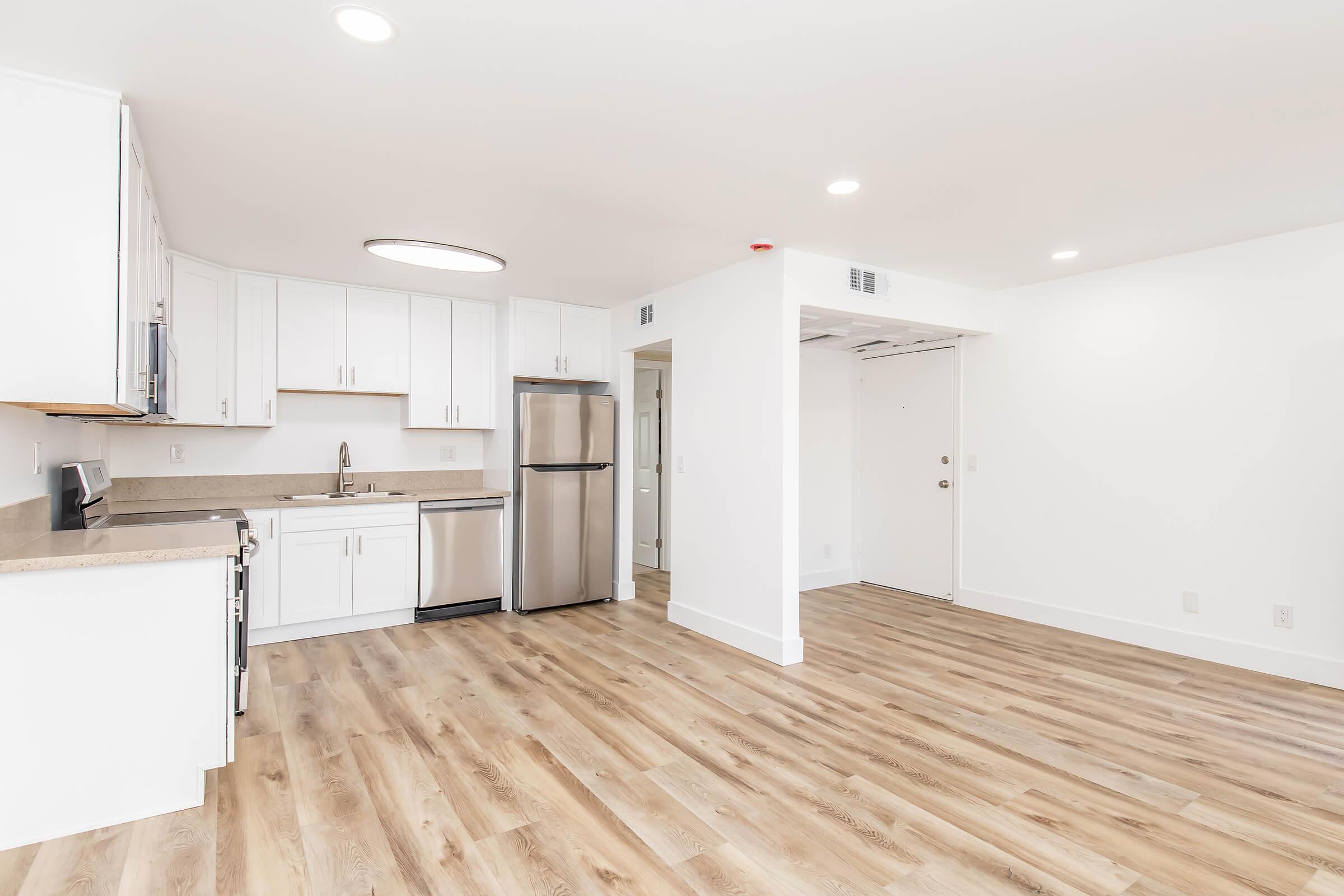 Modern kitchen and dining area with white cabinets, stainless steel appliances, and a spacious layout. The kitchen features a large counter and sink, while the adjacent area has light wood flooring and minimal furnishings, creating an open and bright atmosphere.