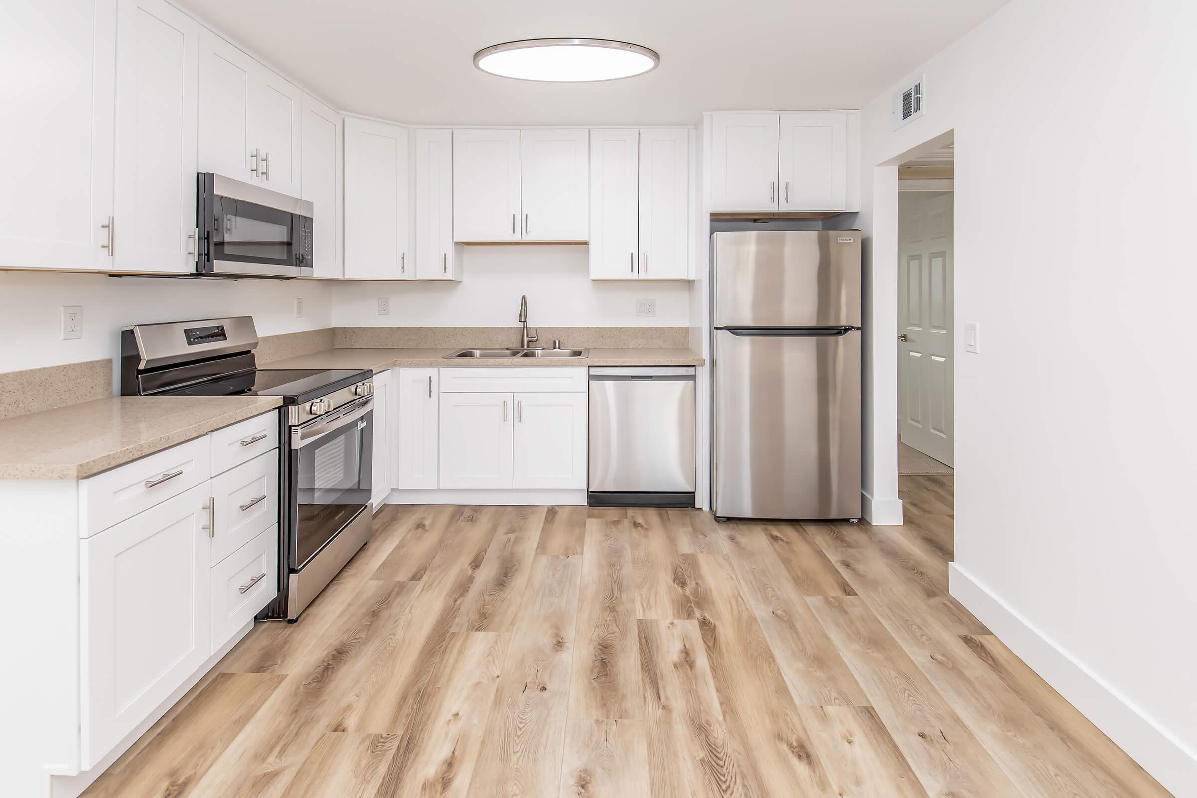 Modern kitchen featuring white cabinets, stainless steel appliances including a microwave, oven, dishwasher, and refrigerator. The countertops are light-colored with a natural finish, and the floor is a light wood laminate. A circular ceiling light provides bright illumination.