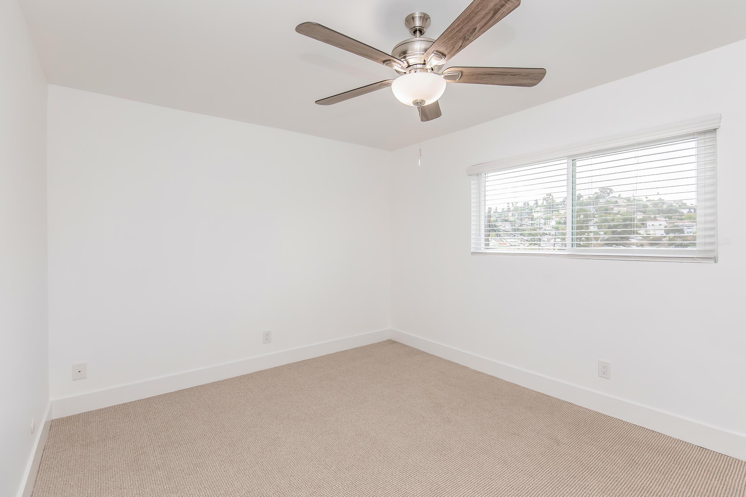 A simple, empty room with light-colored walls and a ceiling fan. There is a window with horizontal blinds that lets in natural light, and the floor is covered with beige carpet. The room appears bright and spacious, ideal for various uses.
