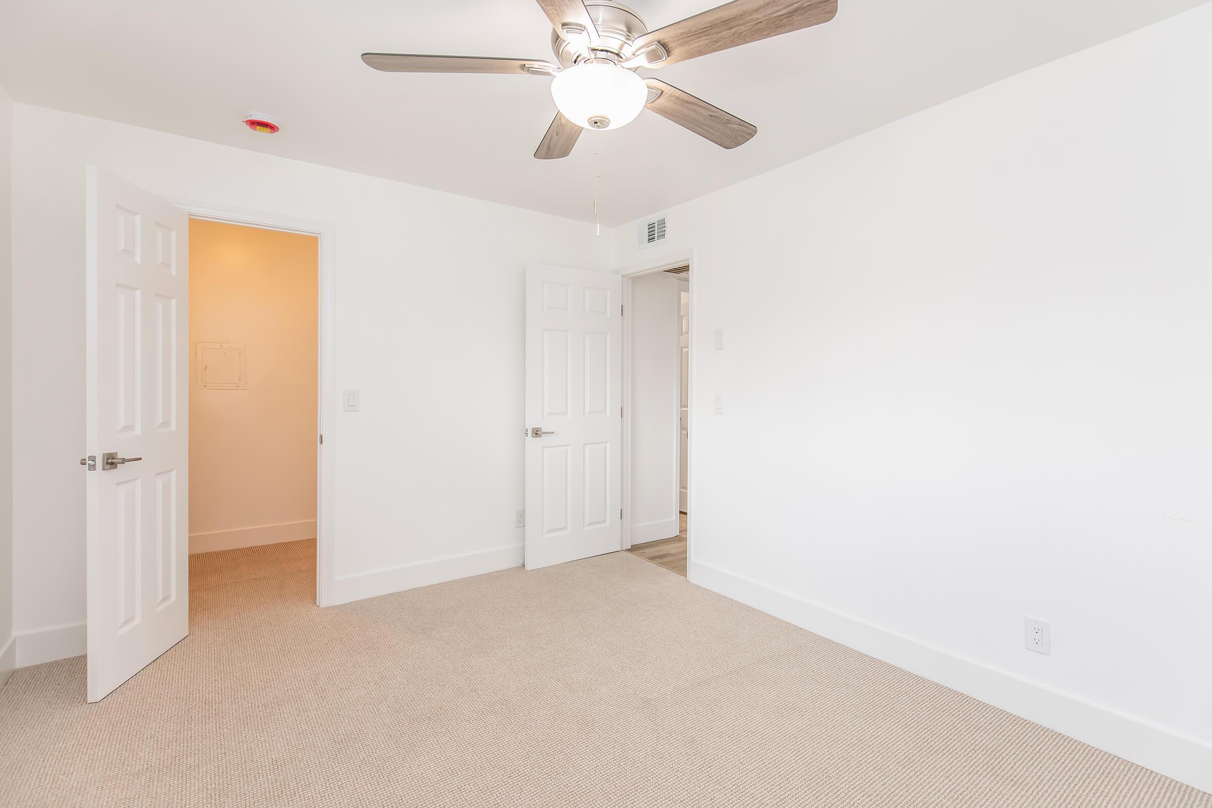 A spacious, brightly lit room featuring a ceiling fan, beige carpet, and white walls. Two doors are visible: one leading to an adjacent room and the other possibly to a closet or bathroom. The room has a simple, modern design with minimal furnishings.