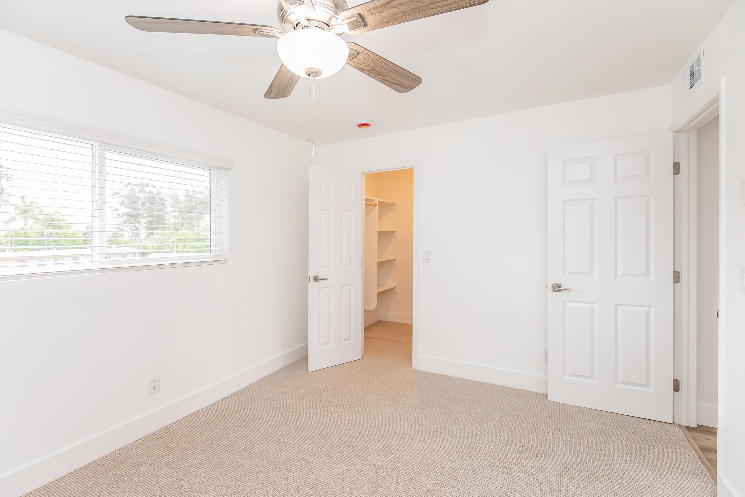 A bright, unfurnished bedroom featuring a ceiling fan, white walls, and a light-colored carpet. One door leads to a closet, while another door opens to an adjacent space. A window on the left allows natural light to enter, creating a spacious and airy atmosphere.
