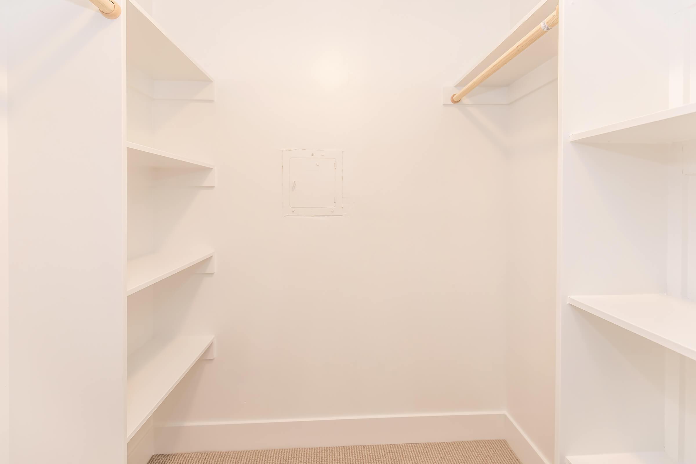 Empty walk-in closet featuring white walls, a beige carpet, two white shelving units on the left, and a wooden hanging rod on the right. The room is well-lit, creating a clean and spacious atmosphere.