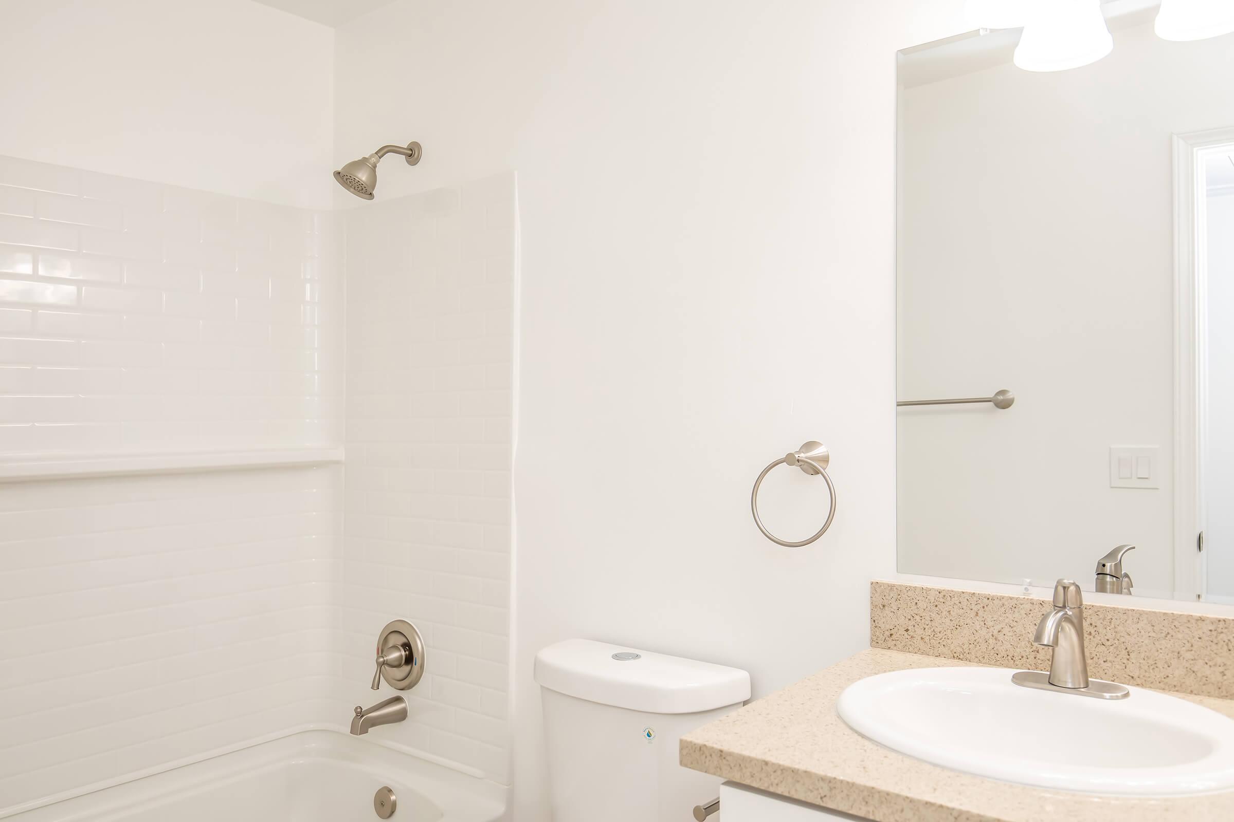 A clean and modern bathroom featuring a white tiled shower and bathtub, a granite countertop with a round sink, a chrome faucet, a simple wall-mounted mirror, and a toilet nearby. The walls are painted white, creating a bright and spacious ambiance.