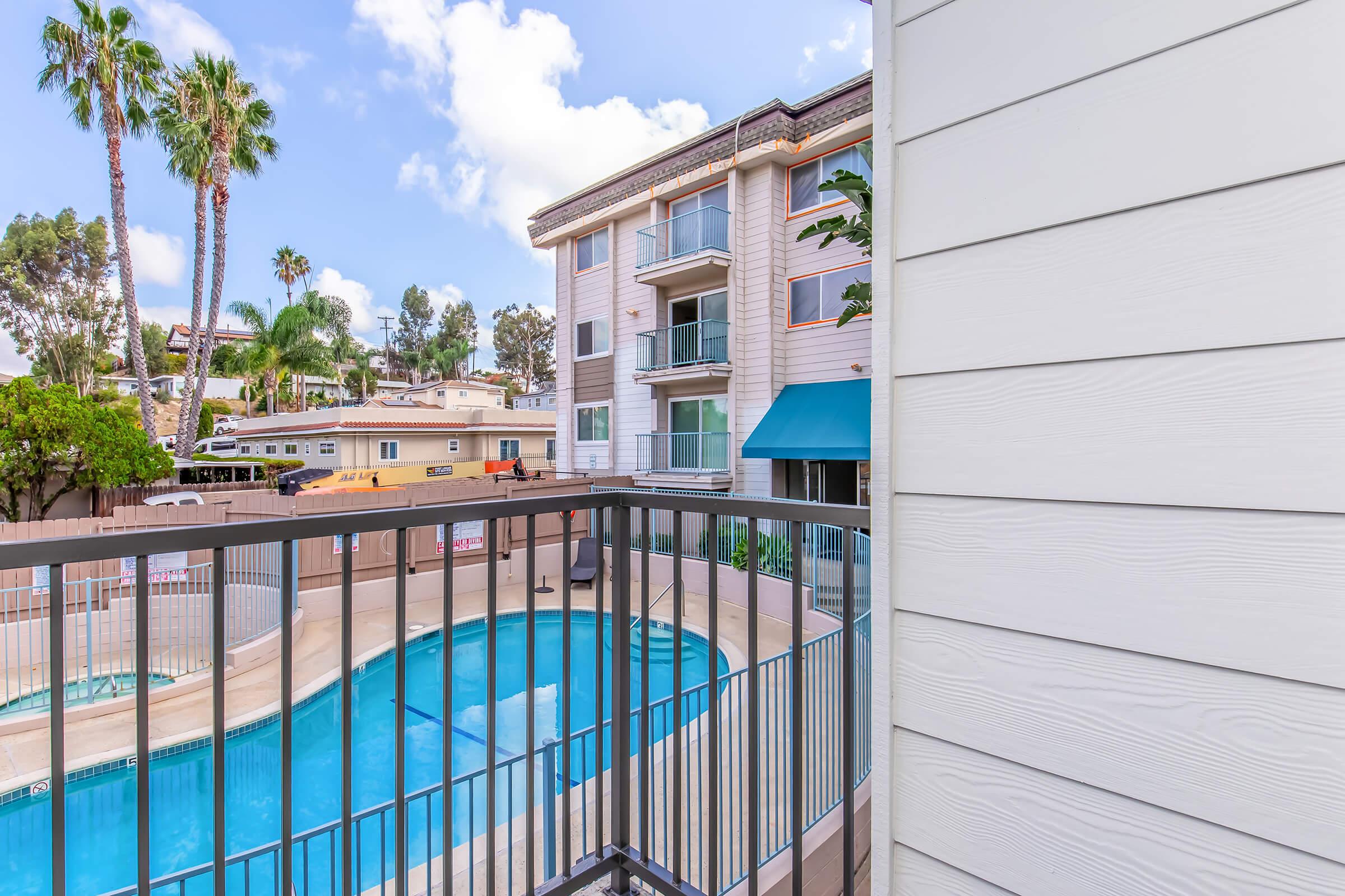 View from a balcony overlooking a swimming pool surrounded by palm trees and residential buildings. The clear blue sky adds to the inviting atmosphere, while the structure's architecture suggests a warm, sunny location, ideal for relaxation and leisure.