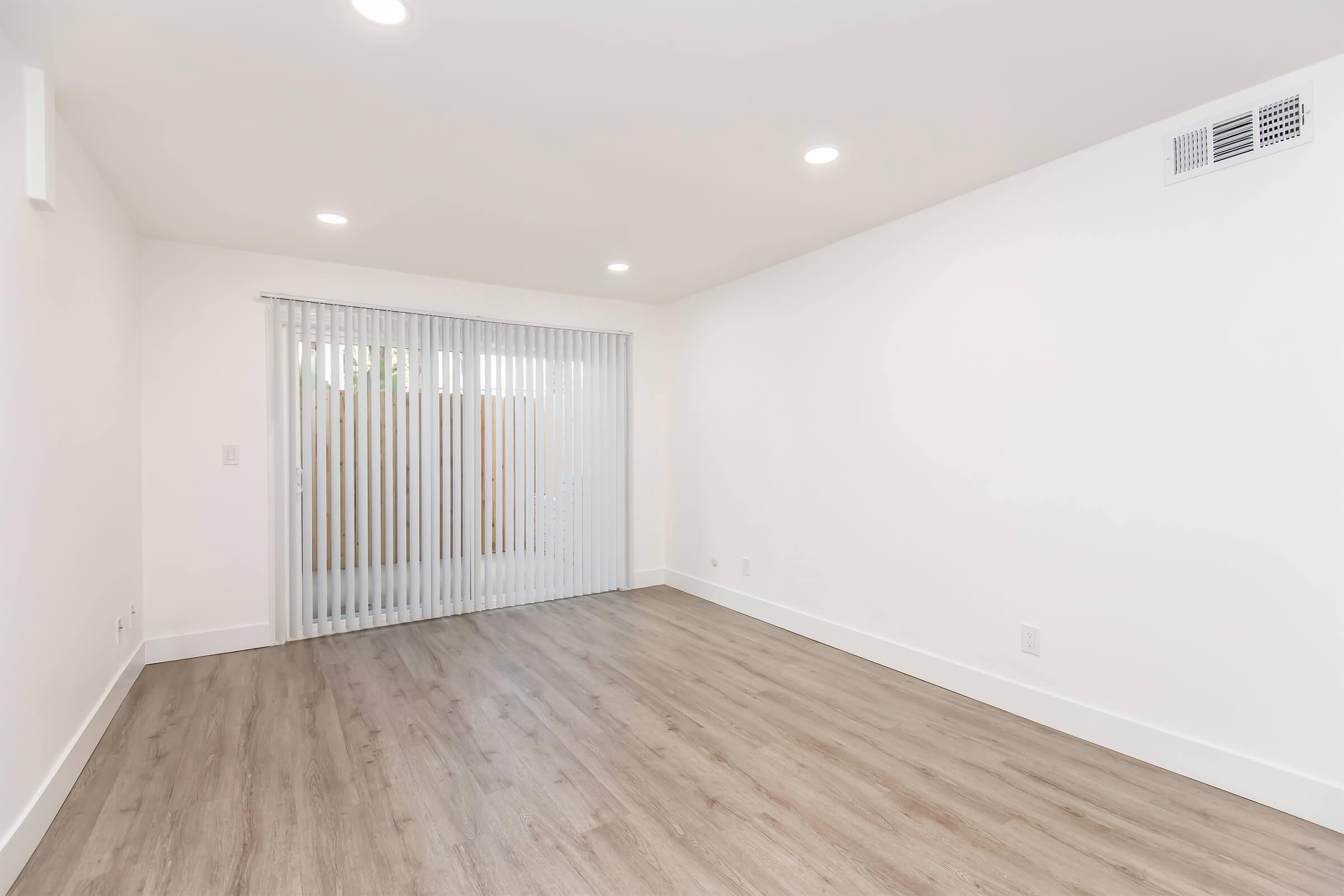 A bright, empty room with light-colored walls and laminate flooring. Large sliding glass doors covered with vertical blinds let in natural light, leading to an outdoor area. The ceiling is equipped with recessed lighting, creating a clean and modern atmosphere.