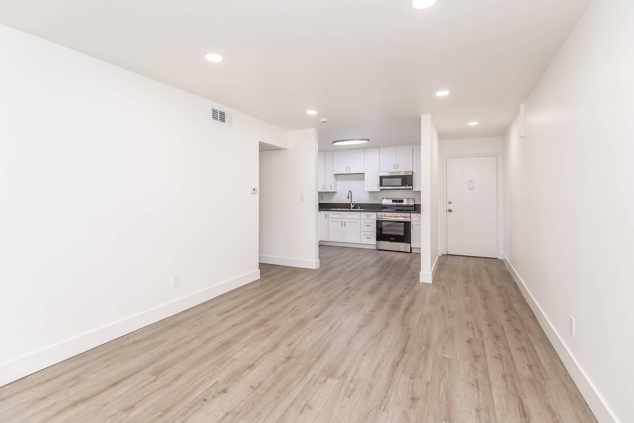 Bright, modern apartment interior featuring an open layout. Light-colored walls, hardwood-style flooring, and recessed lighting create a spacious feel. The kitchen is visible in the background with white cabinetry and stainless steel appliances. A doorway leads to the exterior.