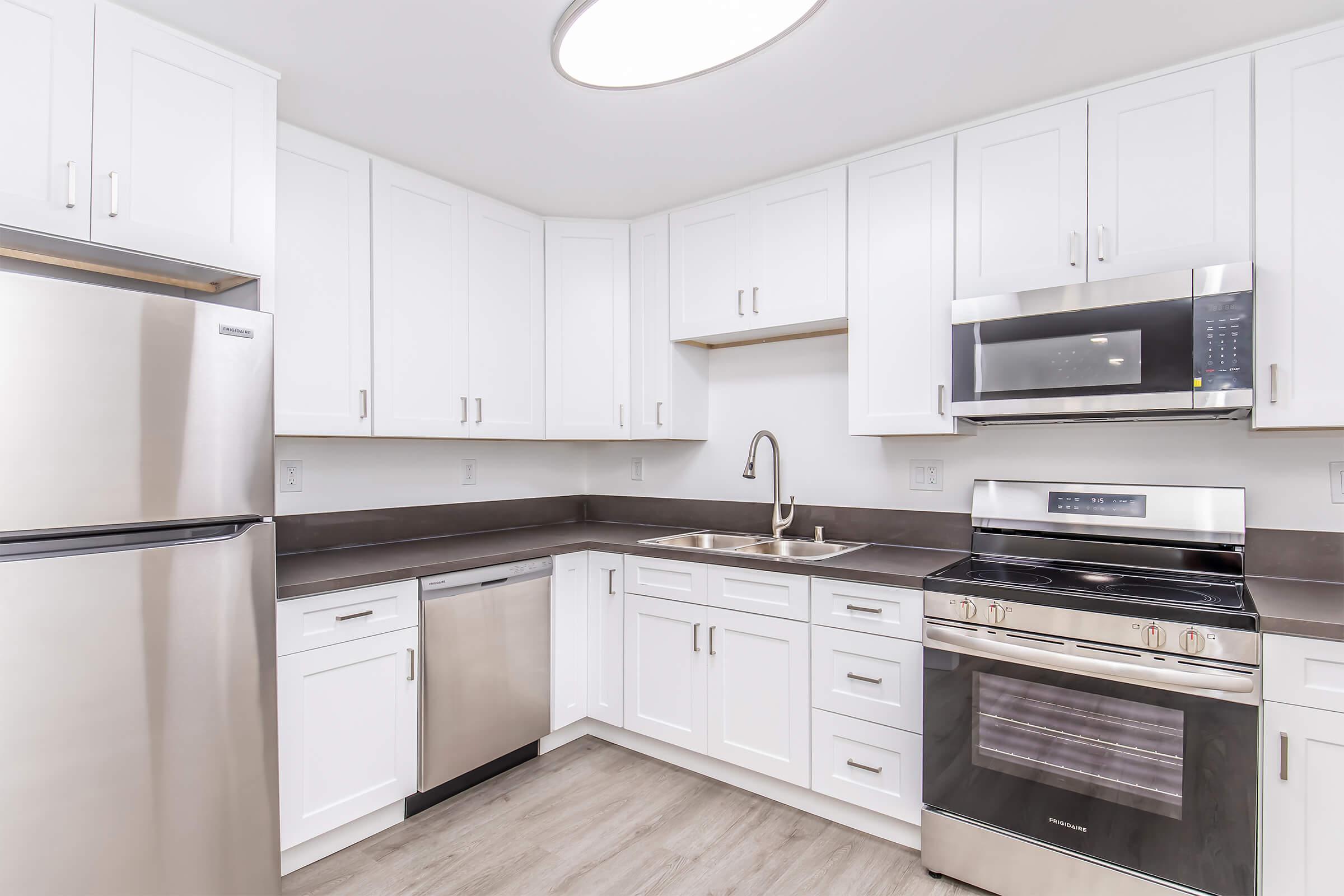Modern kitchen featuring stainless steel appliances, including a refrigerator, dishwasher, microwave, and oven. The space has white cabinetry, a gray countertop, and a sink. Soft lighting from a ceiling fixture enhances the bright, clean aesthetic of the room.